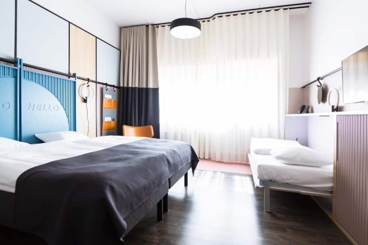 A modern hotel room with a large bed, a smaller bed, a desk, and a chair. There is a window with curtains, a hanging light fixture, and a wall-mounted TV.