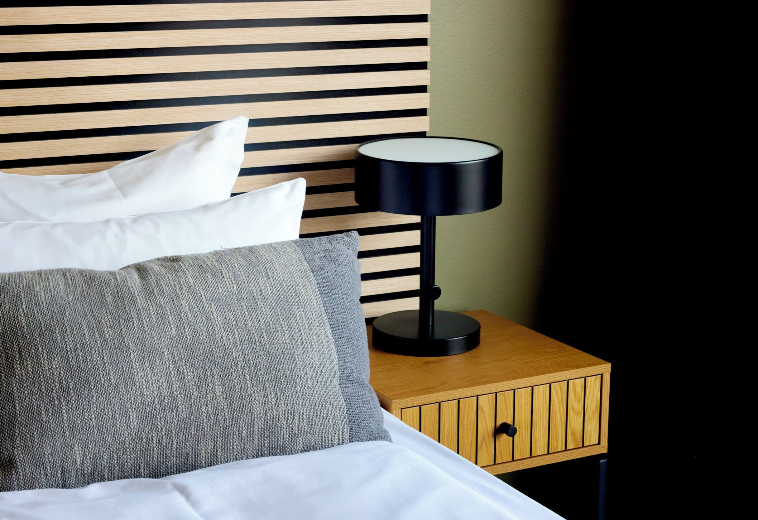 Una cama bien tendida con almohadas, una mesita de noche con lámpara y un cabecero de madera.