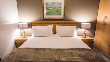 A neatly made bed with white linens and two pillows, flanked by two bedside tables with lamps. A framed landscape painting hangs above the bed.