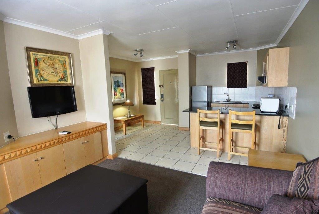 A living room with a flat-screen TV, a sofa, and a coffee table. There is a kitchen area with a refrigerator, sink, and wooden cabinets.