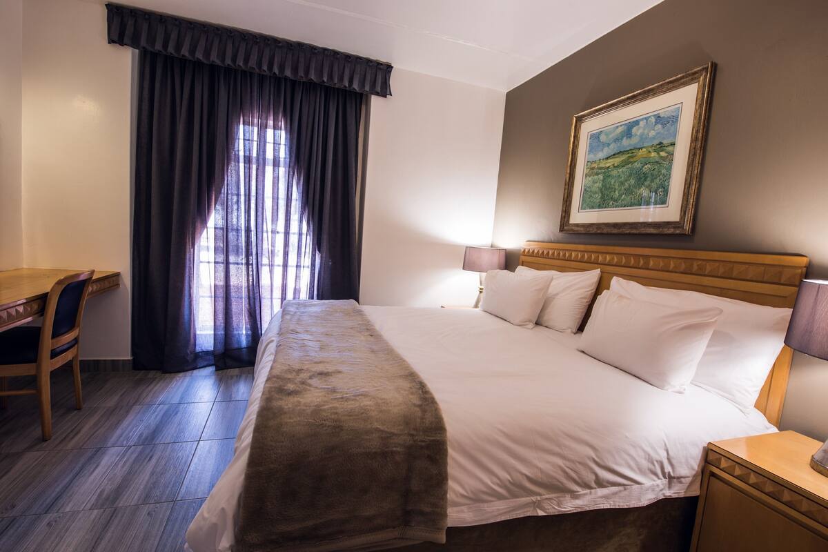 A hotel room with a large bed, a desk, and a chair. There is a painting on the wall and a window with curtains.