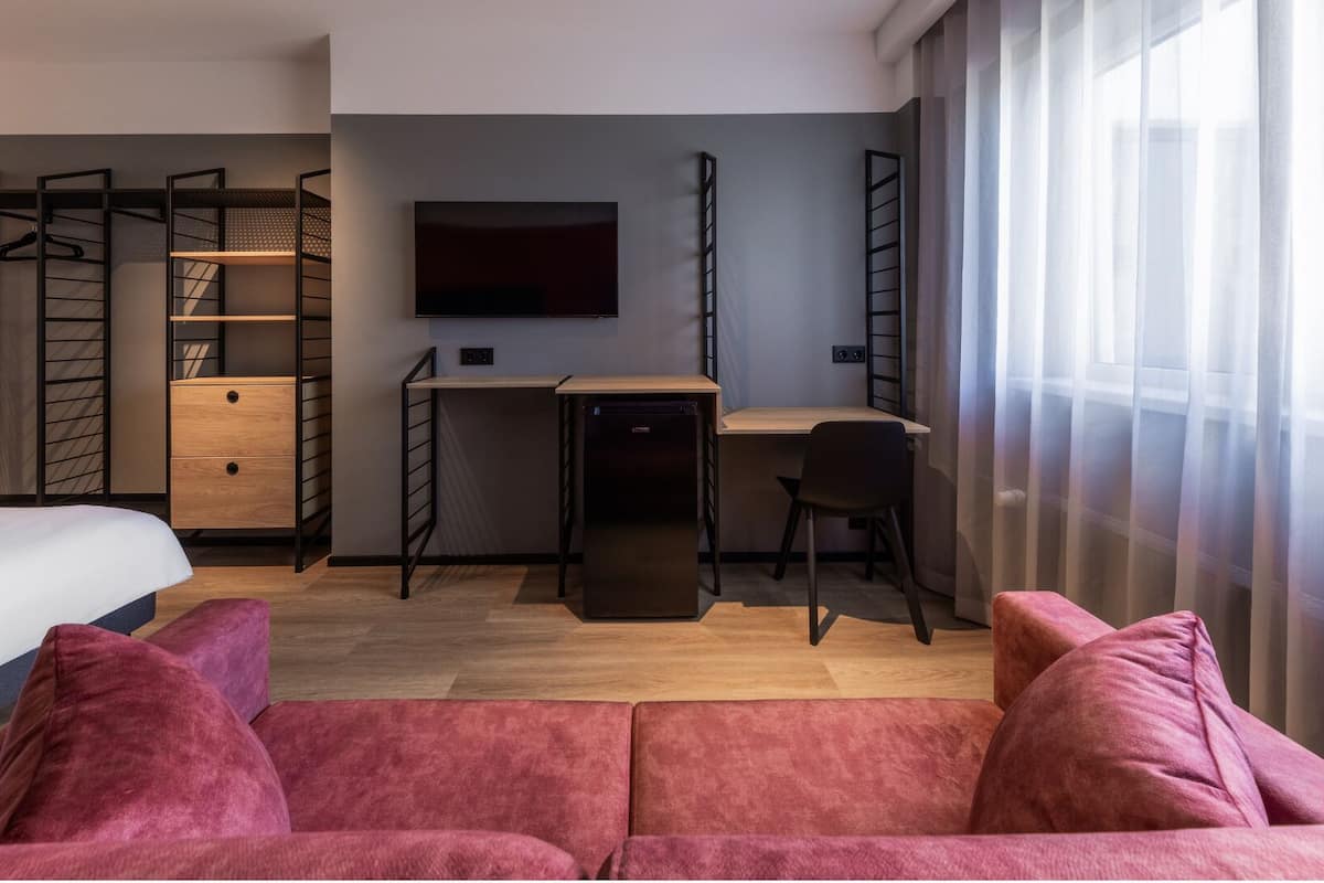 A compact hotel room with a bed, a desk, a chair, a TV, and a sofa.