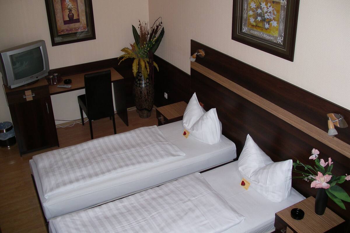 A hotel room with two beds, a desk, a chair, a television, and a vase with flowers.
