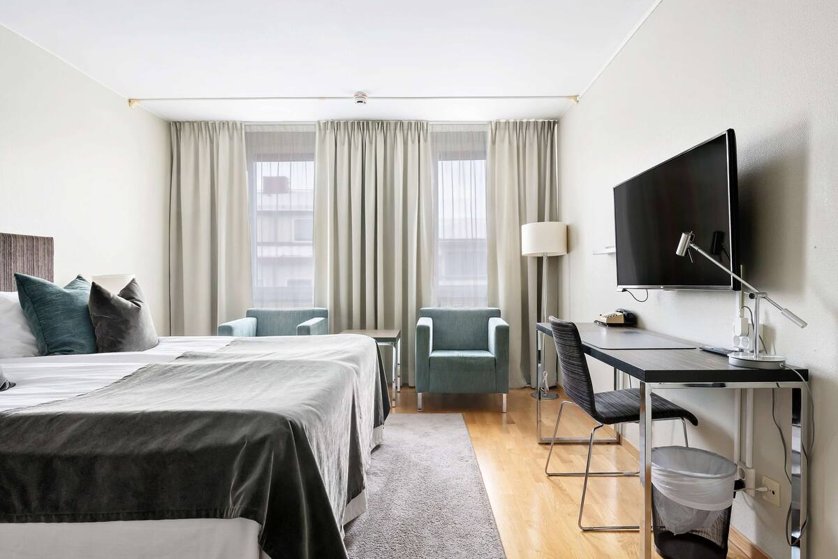 Une chambre d’hôtel avec un lit, un bureau, une chaise, une télévision et une fenêtre avec des rideaux.