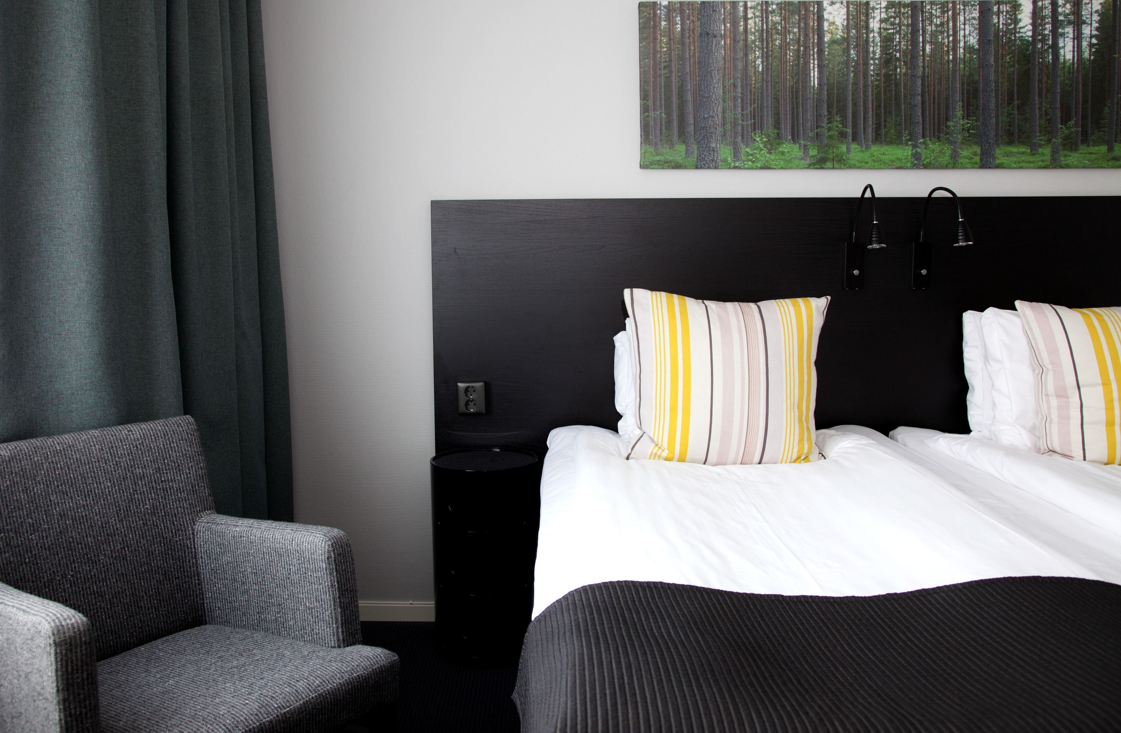 A hotel room with a large bed, two pillows, a bedside table, a chair, and a forest-themed wall art.