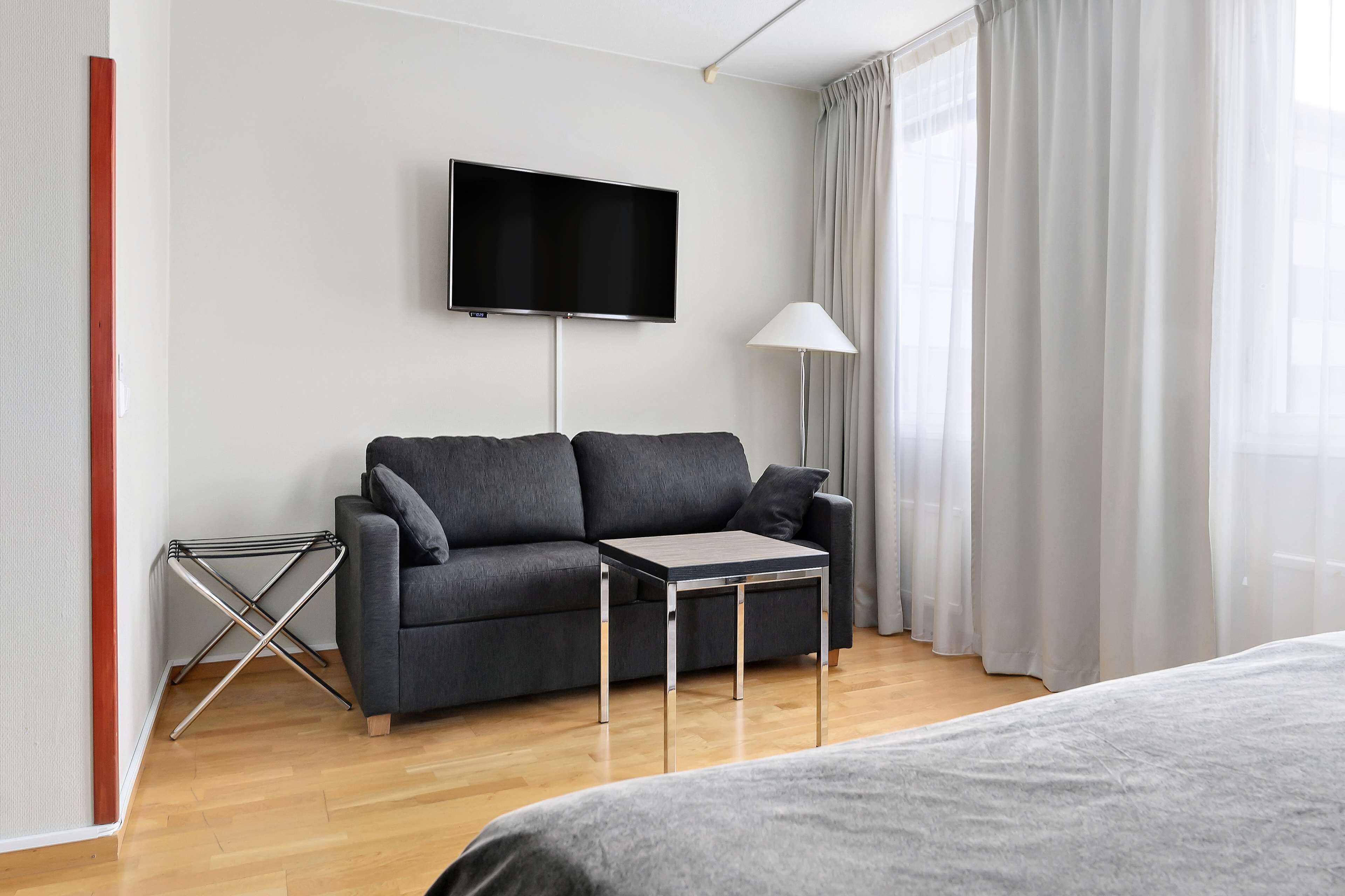 Habitación de hotel moderna con sofá gris, una mesita, televisor montado en la pared y ventana con cortinas.
