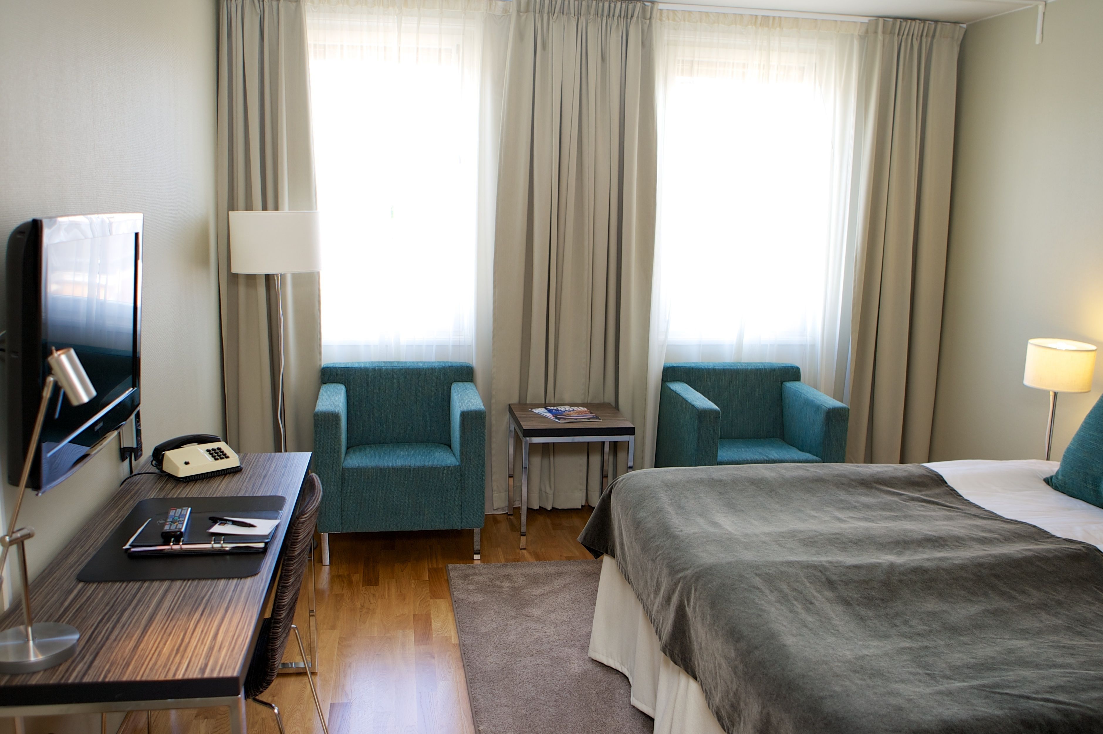 A hotel room with a bed, two armchairs, a desk with a telephone, and a television.