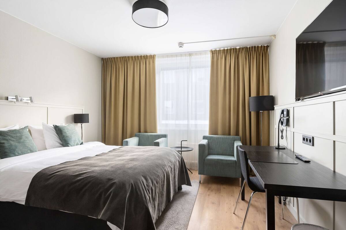 Une chambre d’hôtel avec un grand lit, deux fauteuils, un bureau et une télévision.