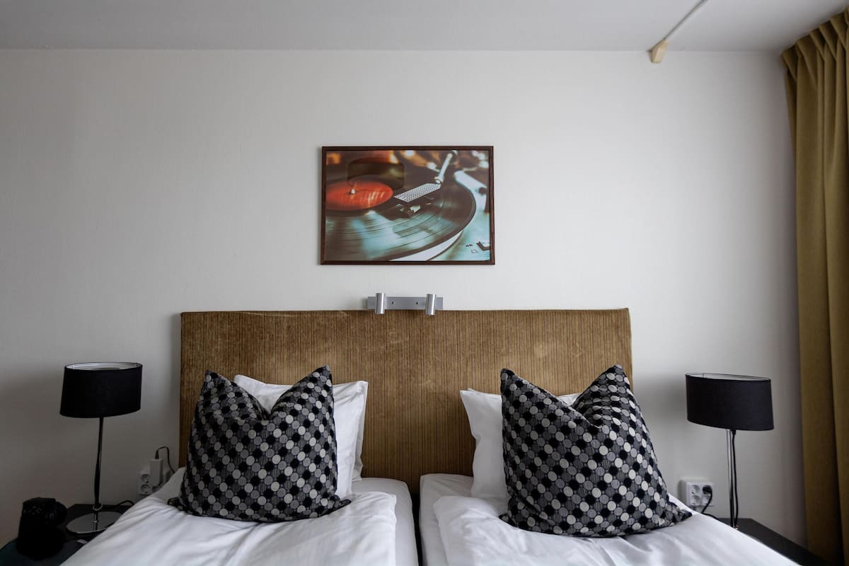 Une chambre d’hôtel avec deux lits, une tête de lit en bois, des coussins à motifs noir et blanc, un tableau accroché au mur et deux lampes de chevet.