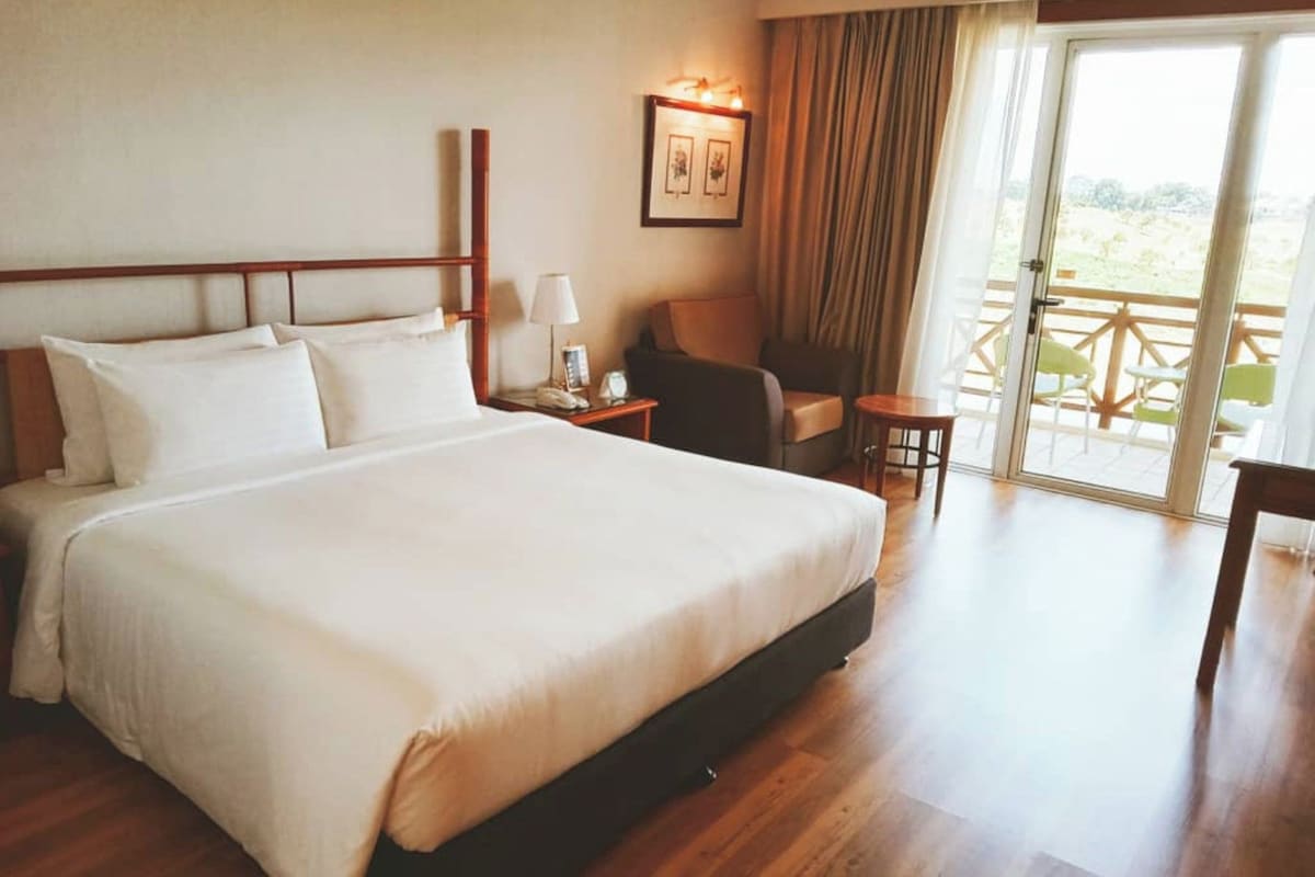 A hotel room with a large bed, a chair, a small table, and a balcony with outdoor furniture.