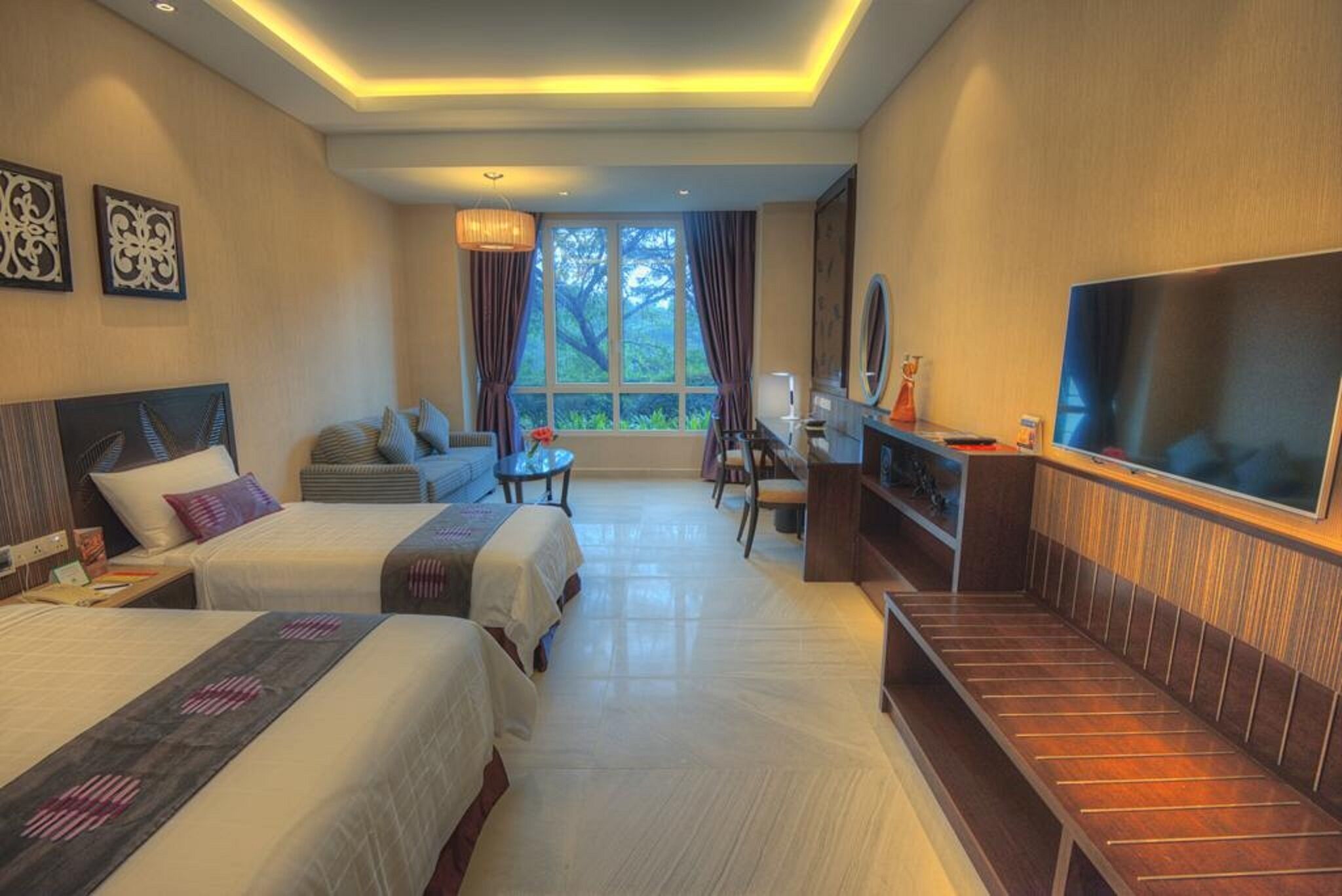 A hotel room with two beds, a sofa, a desk, a TV, and a large window with curtains.