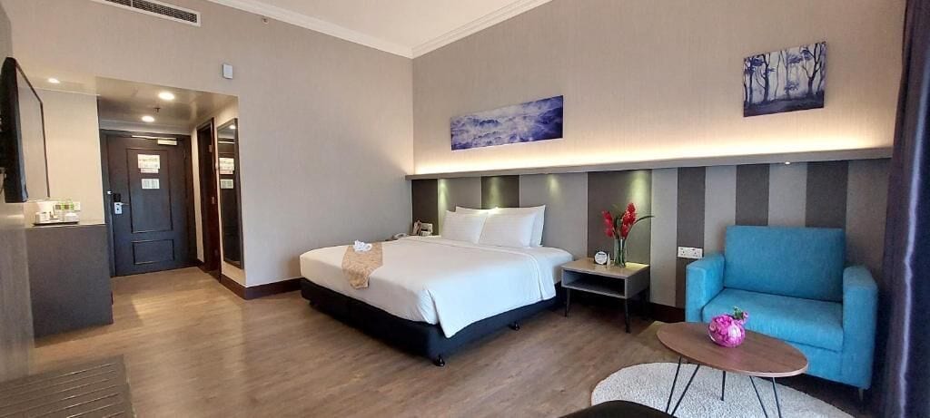 A hotel room with a bed, a blue armchair, a small table, and a painting on the wall.