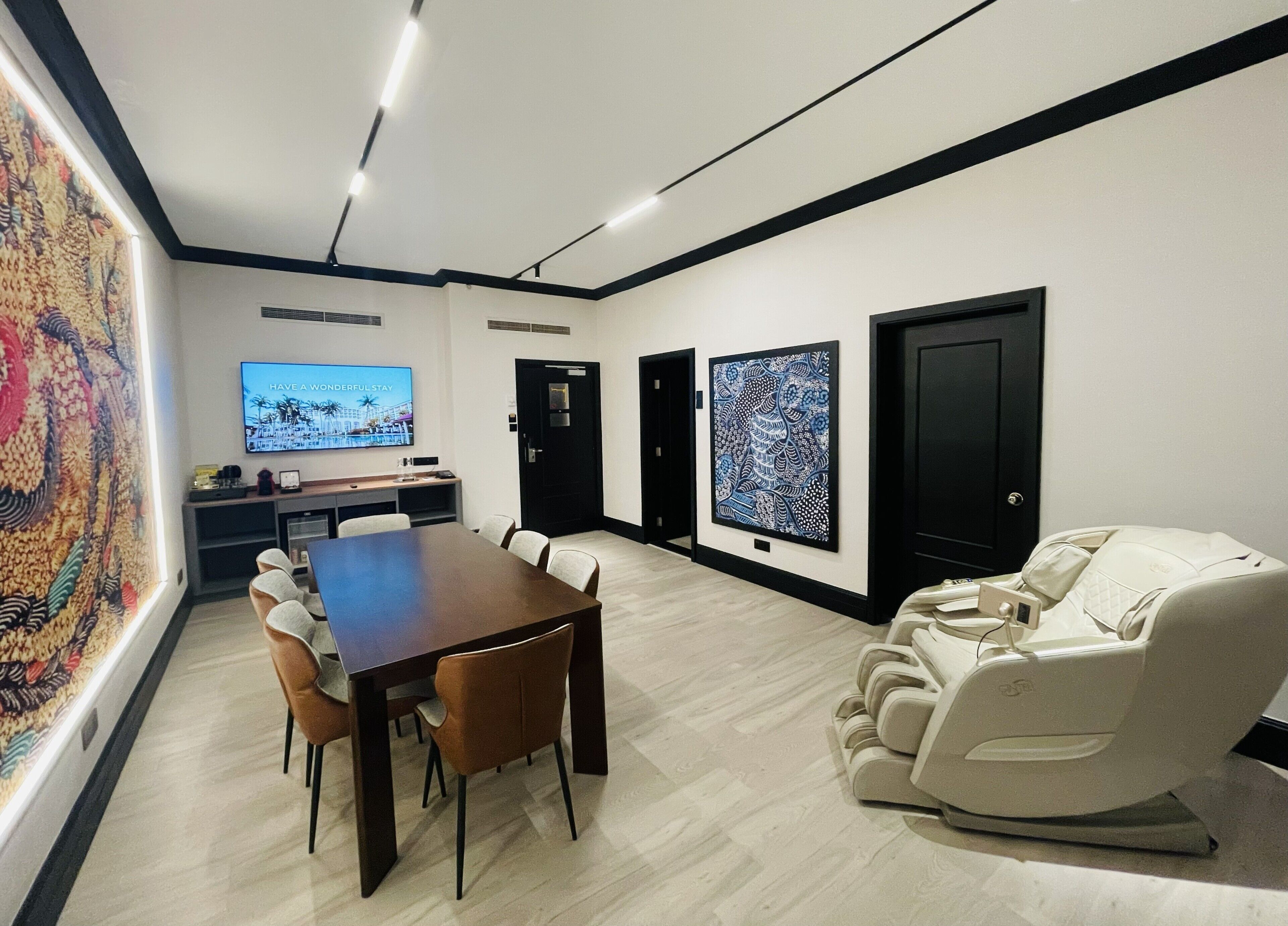 A modern hotel room with a dining area, a massage chair, artwork on the walls, and a television.