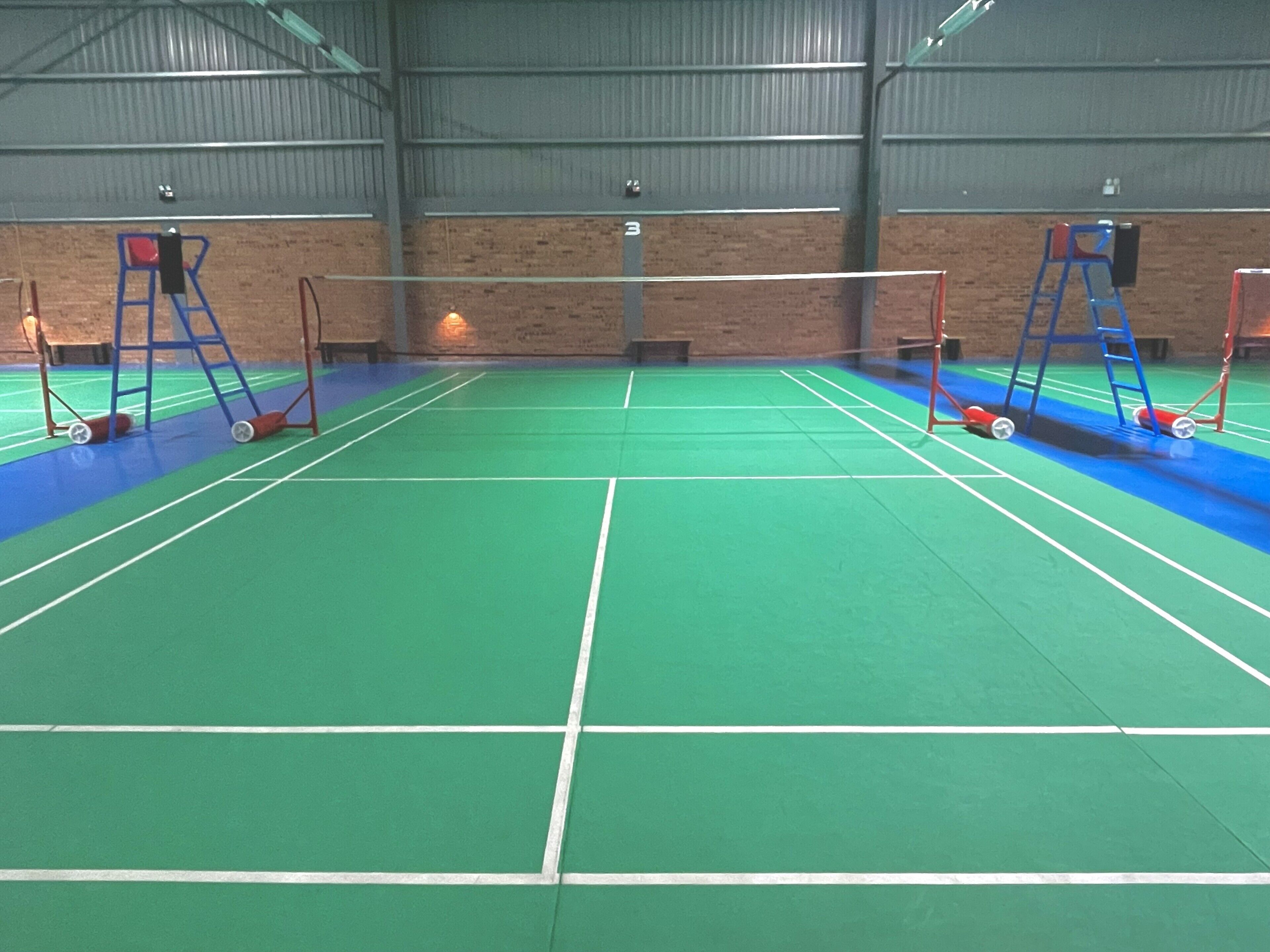 An indoor badminton court with a green floor, white boundary lines, and red and blue equipment.