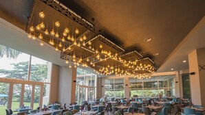 Un comedor amplio con varias mesas y sillas, ventanas grandes que ofrecen vistas al exterior y lámparas colgantes.