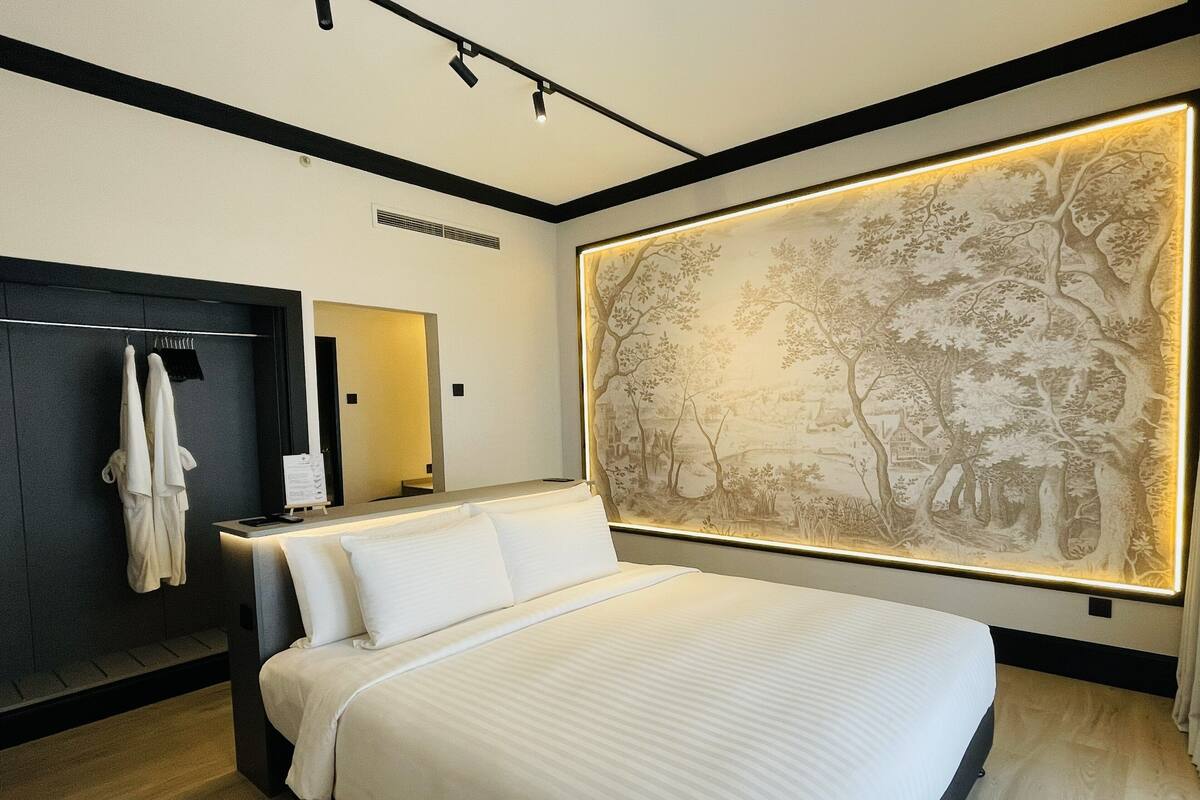 A modern hotel room with a large bed, a dark wooden wardrobe, a mirror, and a wall-mounted artwork depicting a tree scene.