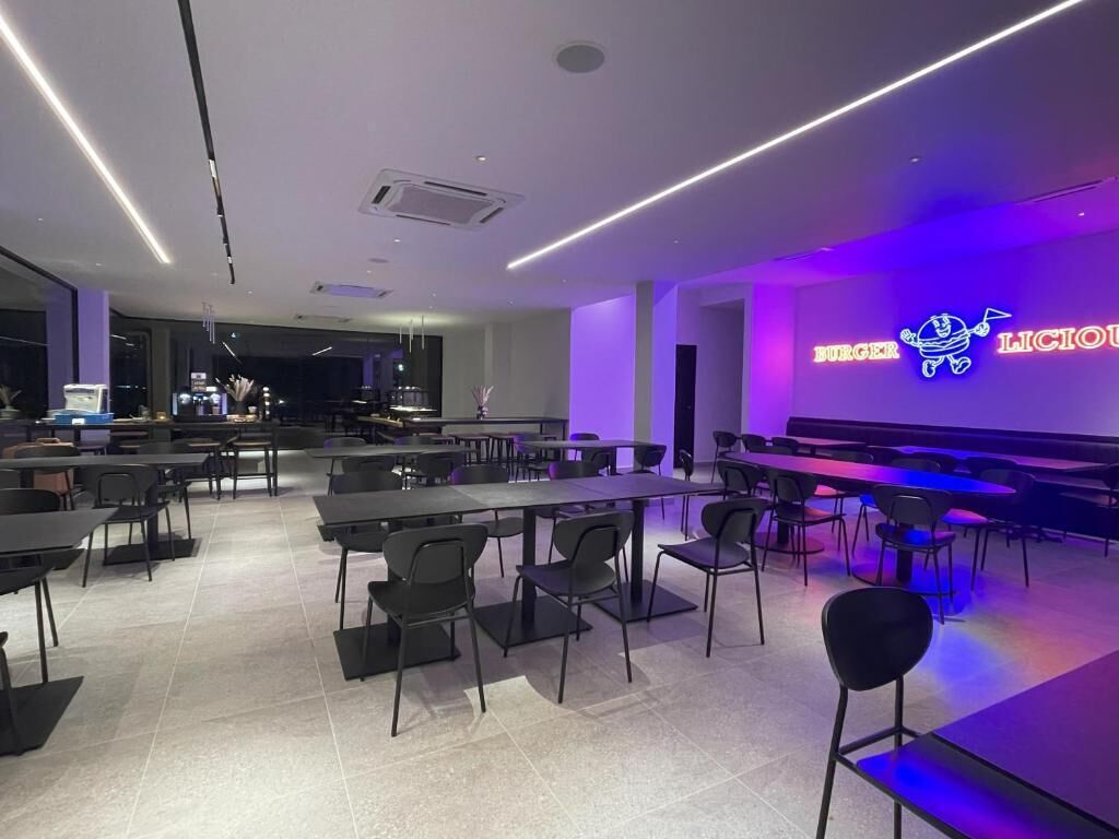 Un comedor moderno con mesas y sillas, un letrero de neón que dice "BURGER LICIOU" y un piano al fondo.