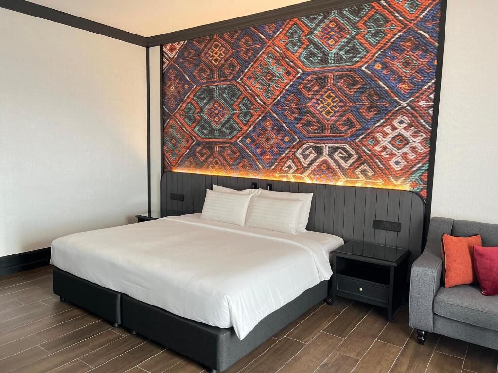 A modern hotel room with a large bed, a grey sofa, and a patterned wall tapestry.