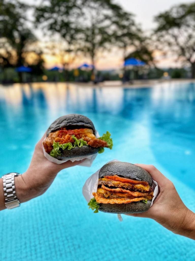 Dos manos sosteniendo hamburguesas sobre una piscina.