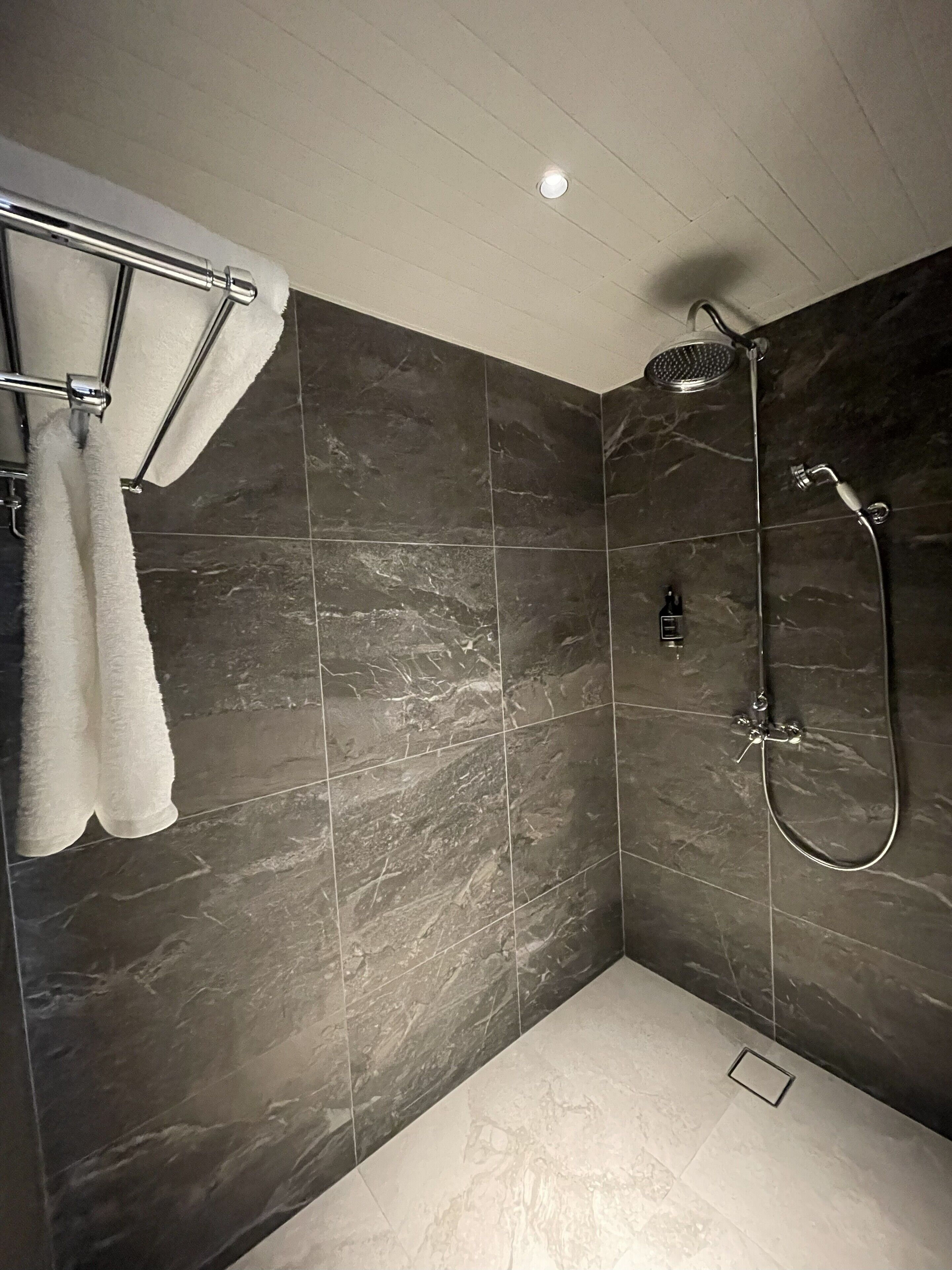 A modern shower area with dark marble tiles, a handheld showerhead, and a towel rack with white towels.