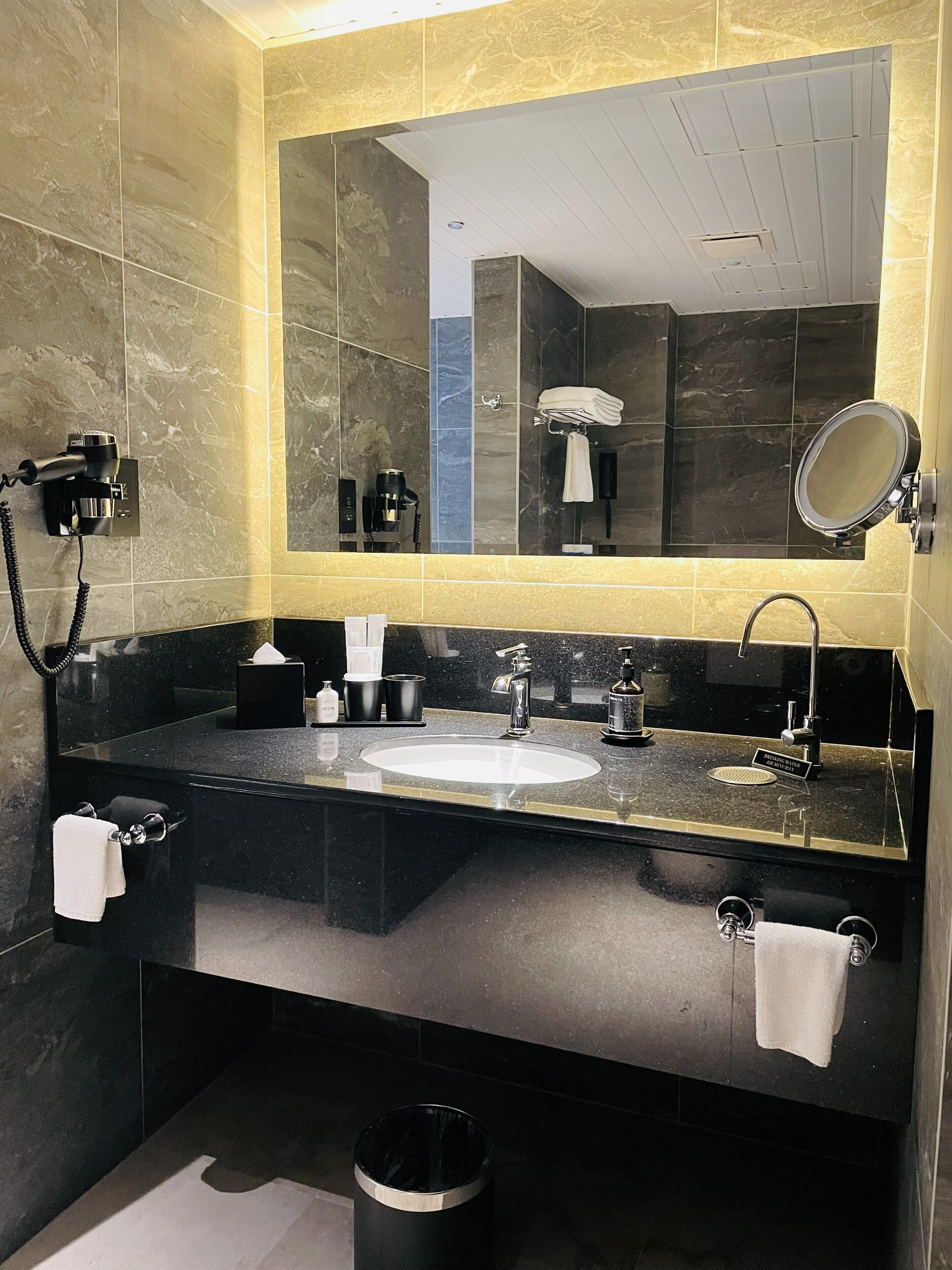 A modern bathroom with a black granite countertop, a white sink, a large mirror, a towel rack, and a trash can.