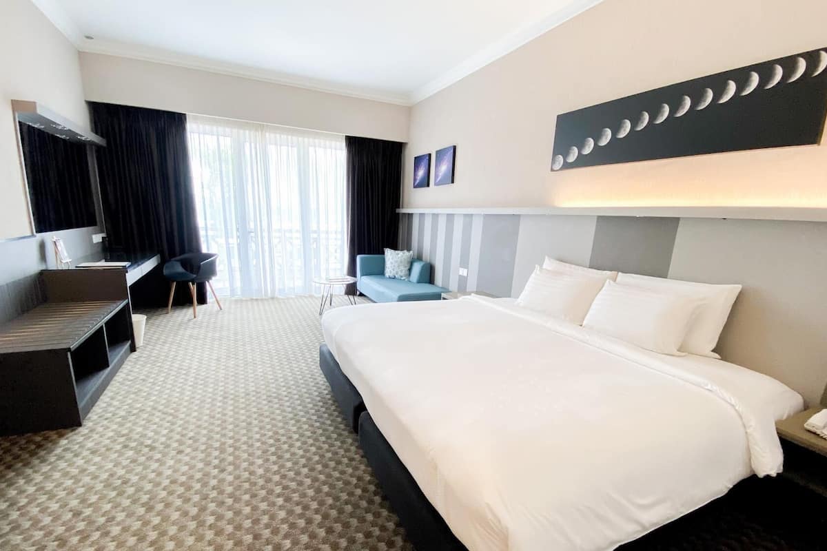 A hotel room with a large bed, a desk, a chair, and a sofa. There is a window with curtains, a wall-mounted phone, and a wall art piece of moons.