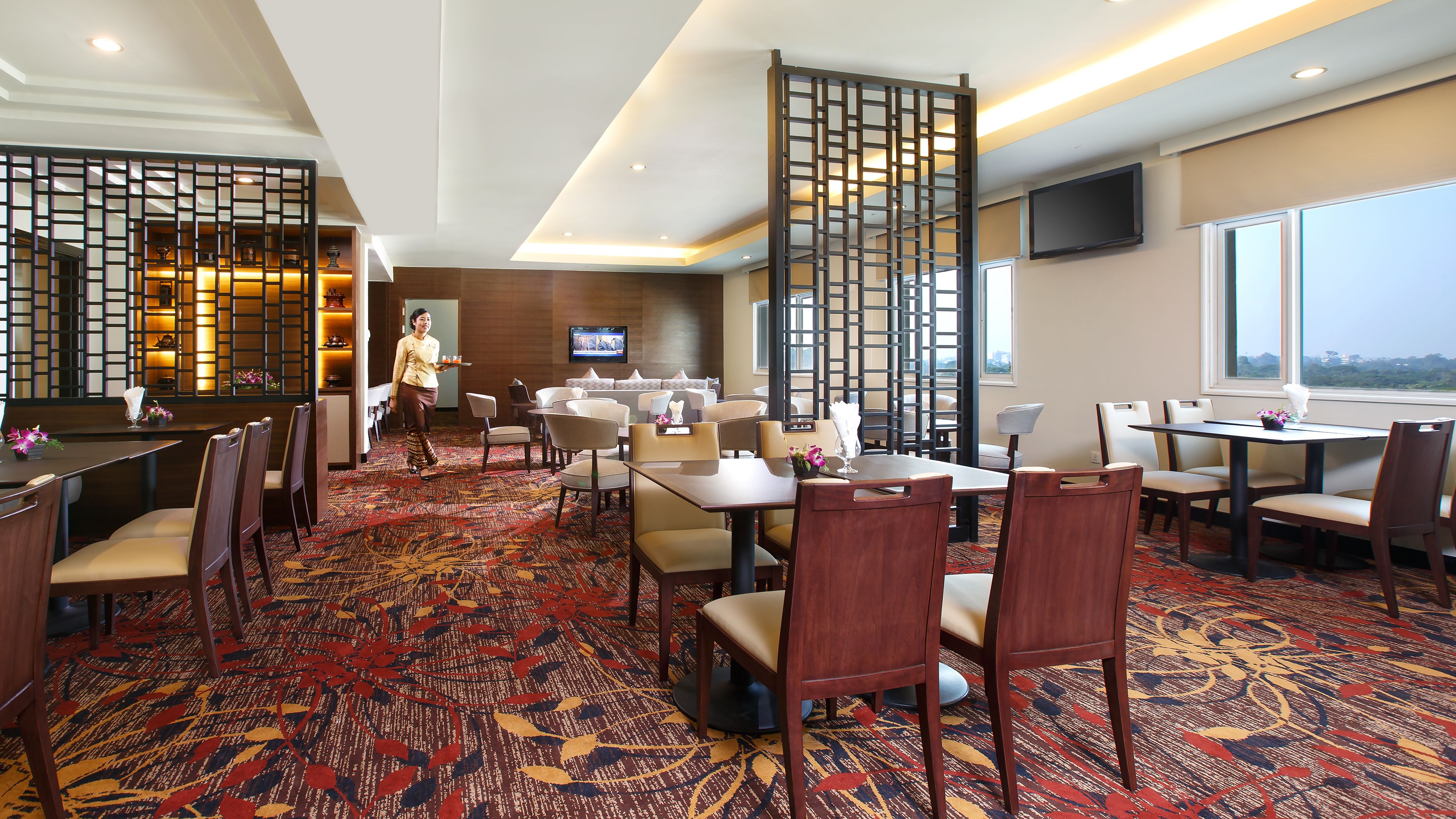 A hotel dining area with multiple tables and chairs, a TV mounted on the wall, and a view of the city through large windows.