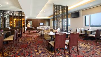 A hotel dining area with multiple tables and chairs, a TV mounted on the wall, and a view of the city through large windows.