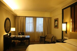Habitación de hotel con cama, escritorio, silla, mesita, lámpara, espejo y ventana con cortinas.