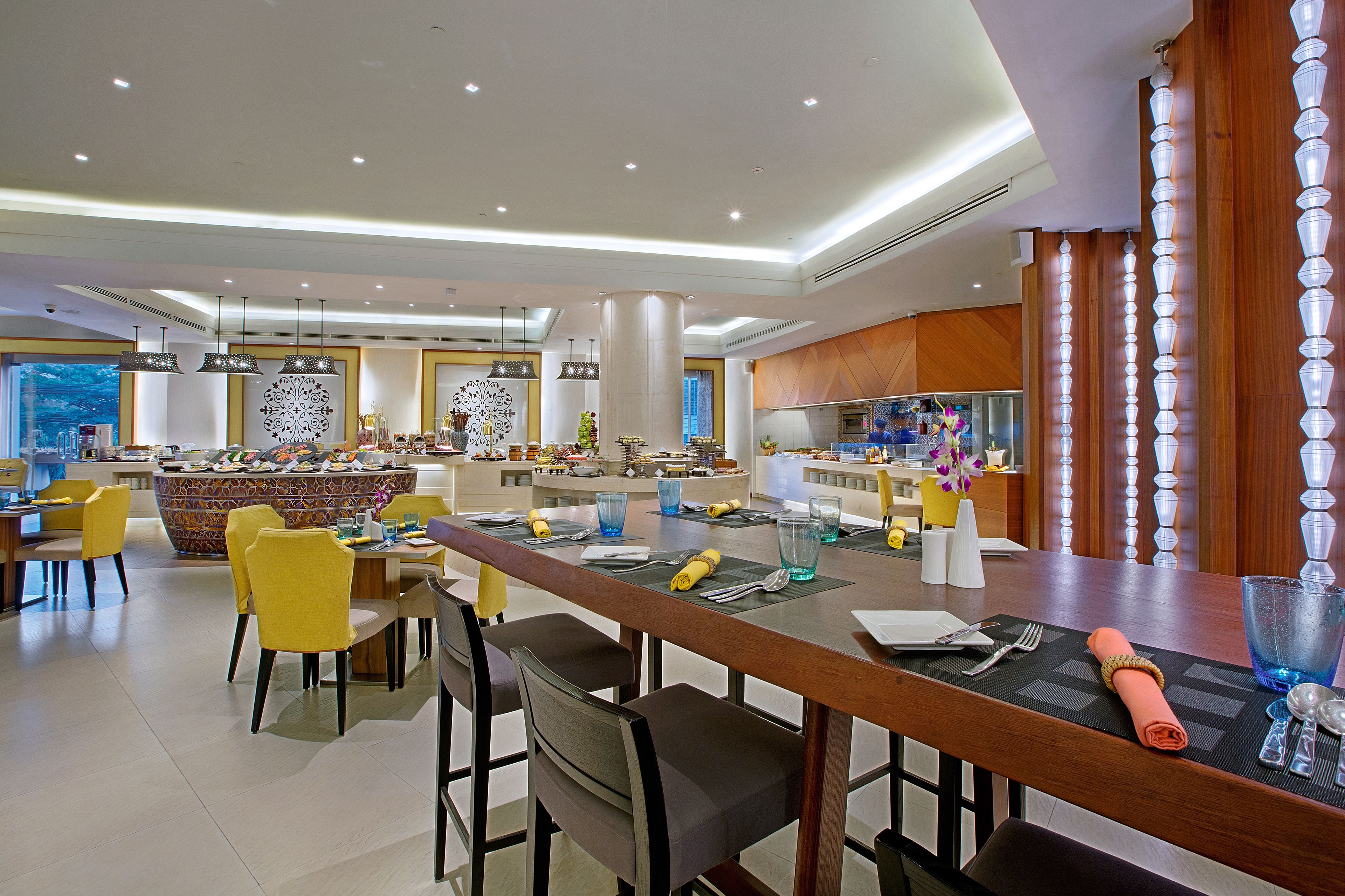 Un comedor moderno de hotel con una larga mesa de madera, sillas amarillas y una isla central con un salpicadero colorido.