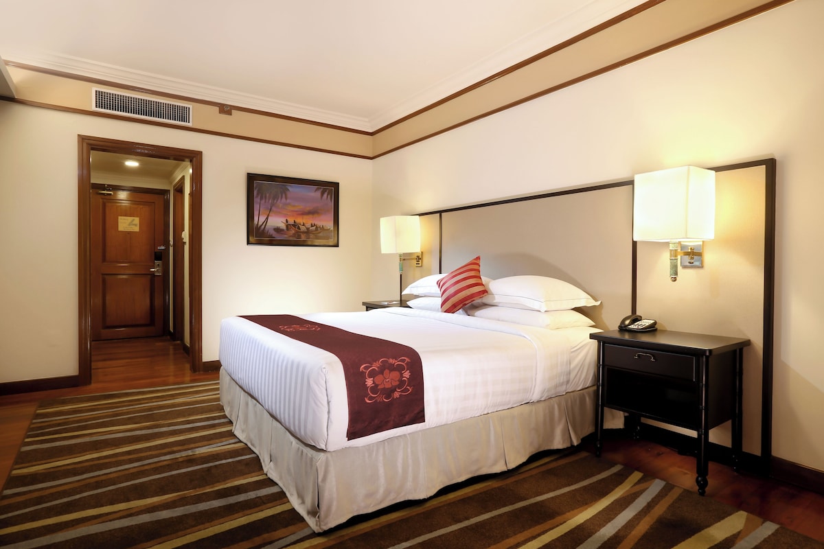 Una habitación de hotel con una cama grande, dos mesitas de noche con lámparas, un cuadro en la pared y una puerta que da a otra habitación.
