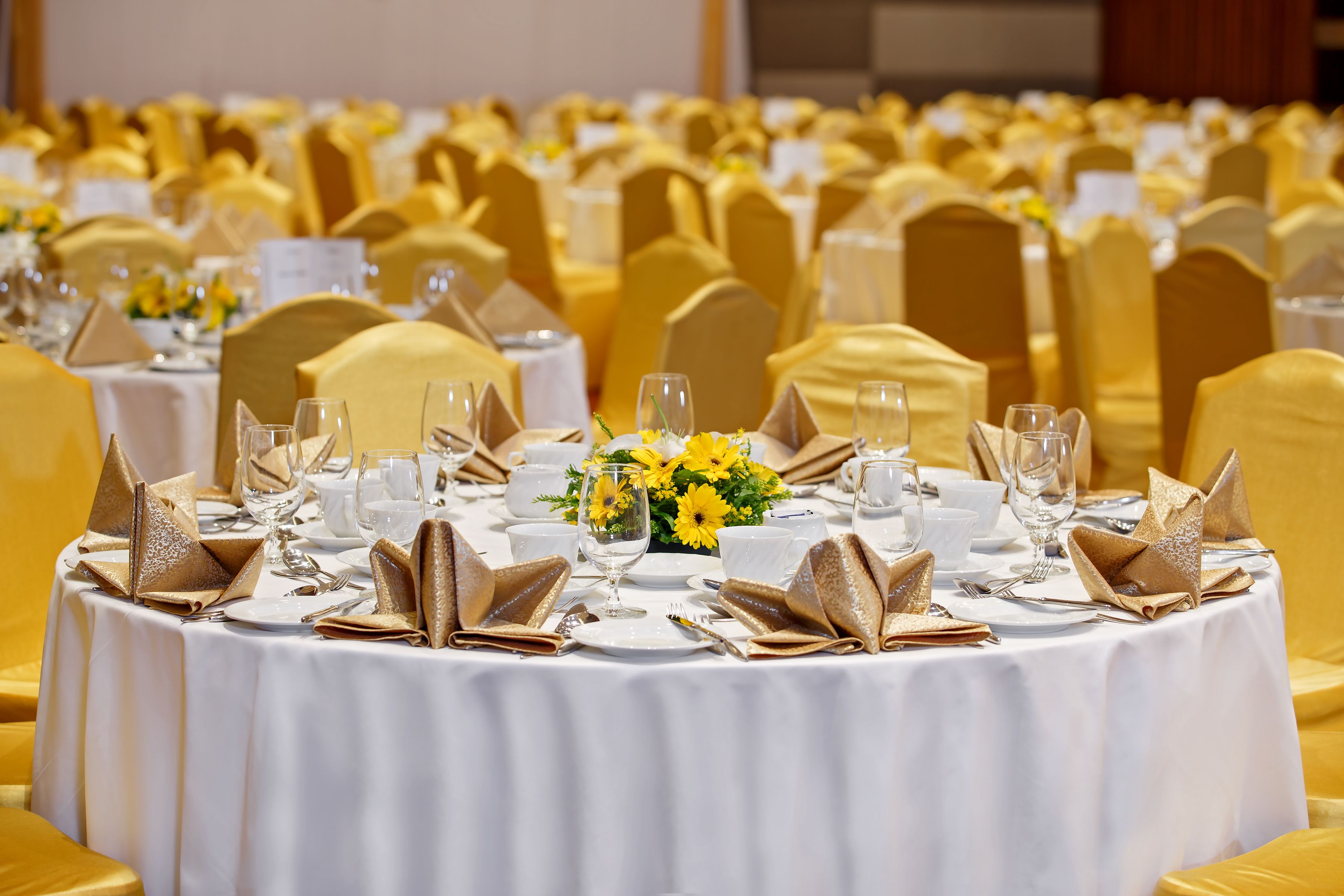A banquet hall with round tables set for a formal event, featuring white tablecloths, gold napkins, and yellow chairs.