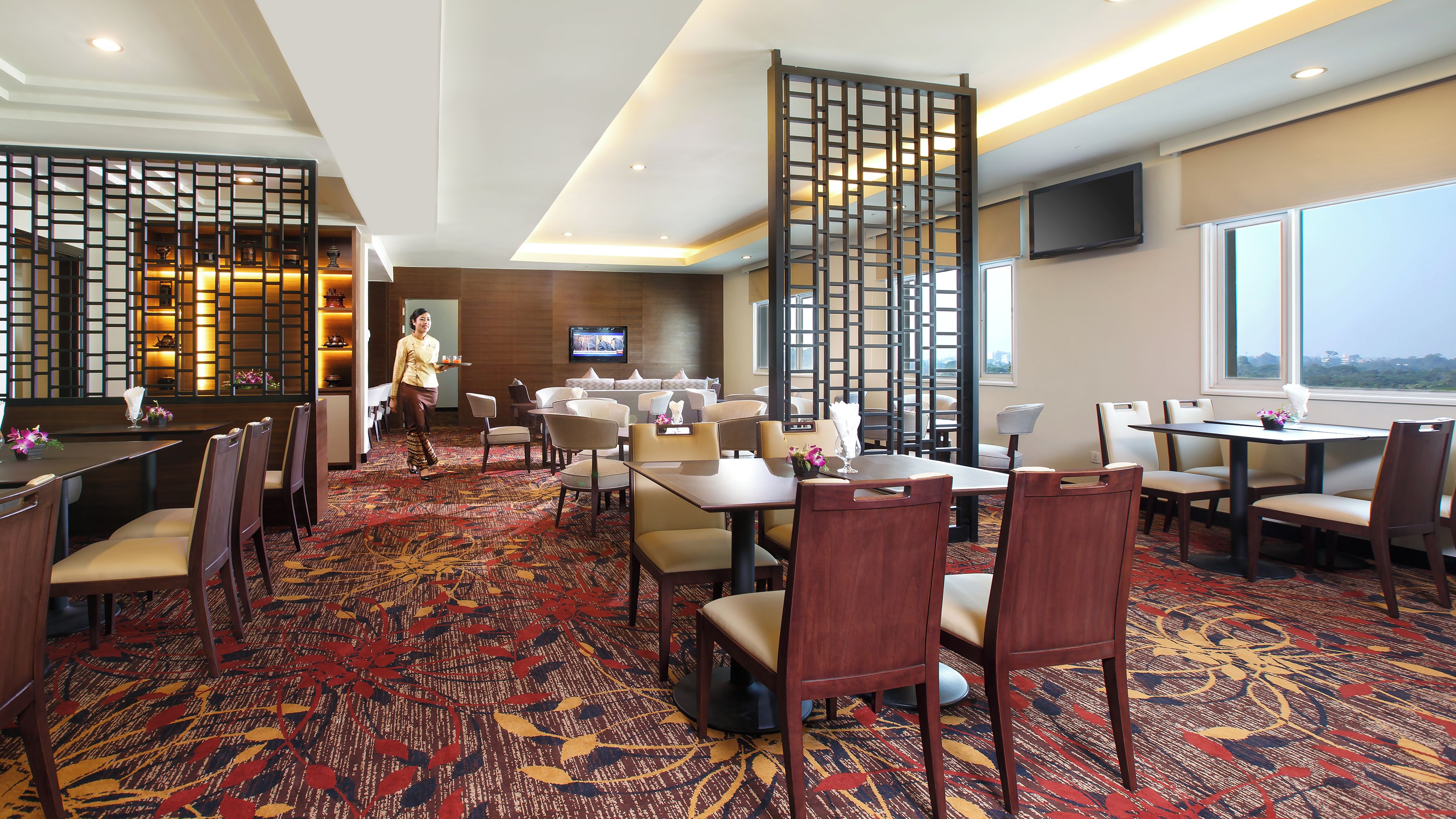 A hotel dining area with multiple tables and chairs, a TV mounted on the wall, and a view of the cityscape through the windows.