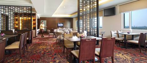 A hotel dining area with multiple tables and chairs, a TV mounted on the wall, and a view of the cityscape through the windows.