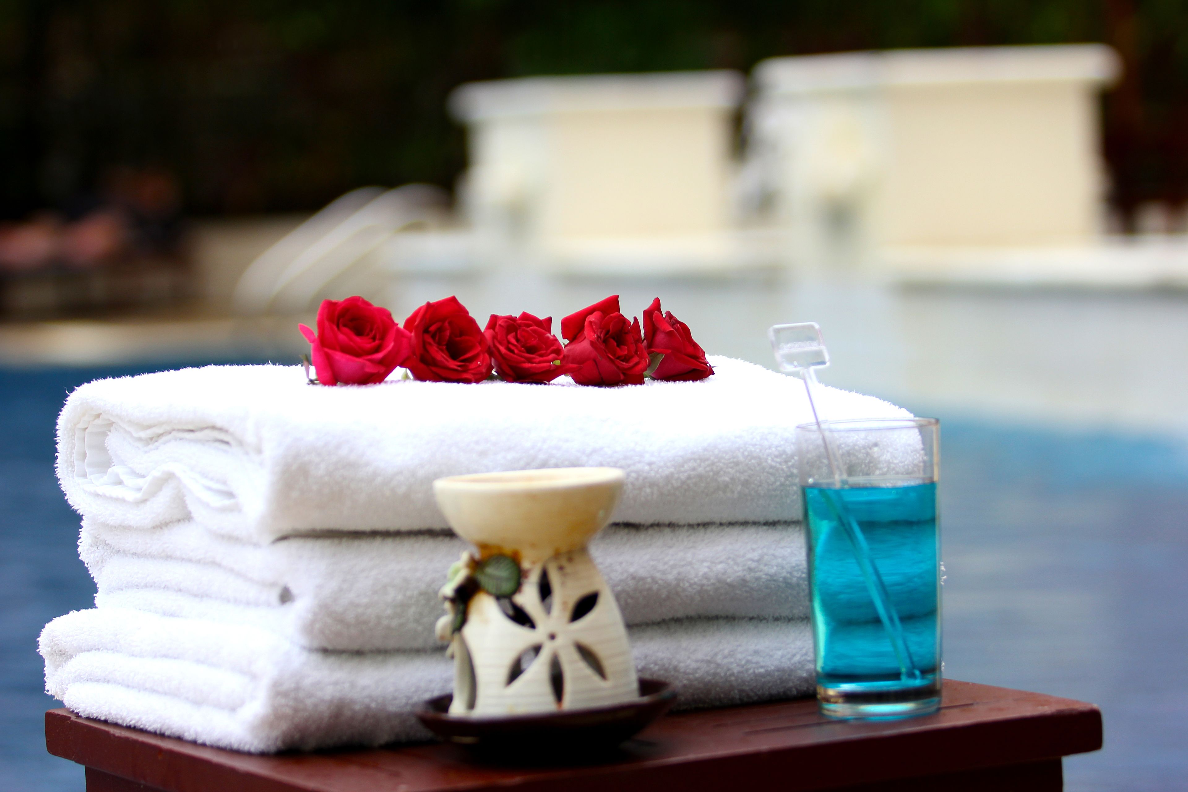 A stack of white towels, a small vase with a flower, and a glass of blue liquid on a wooden table by a pool.