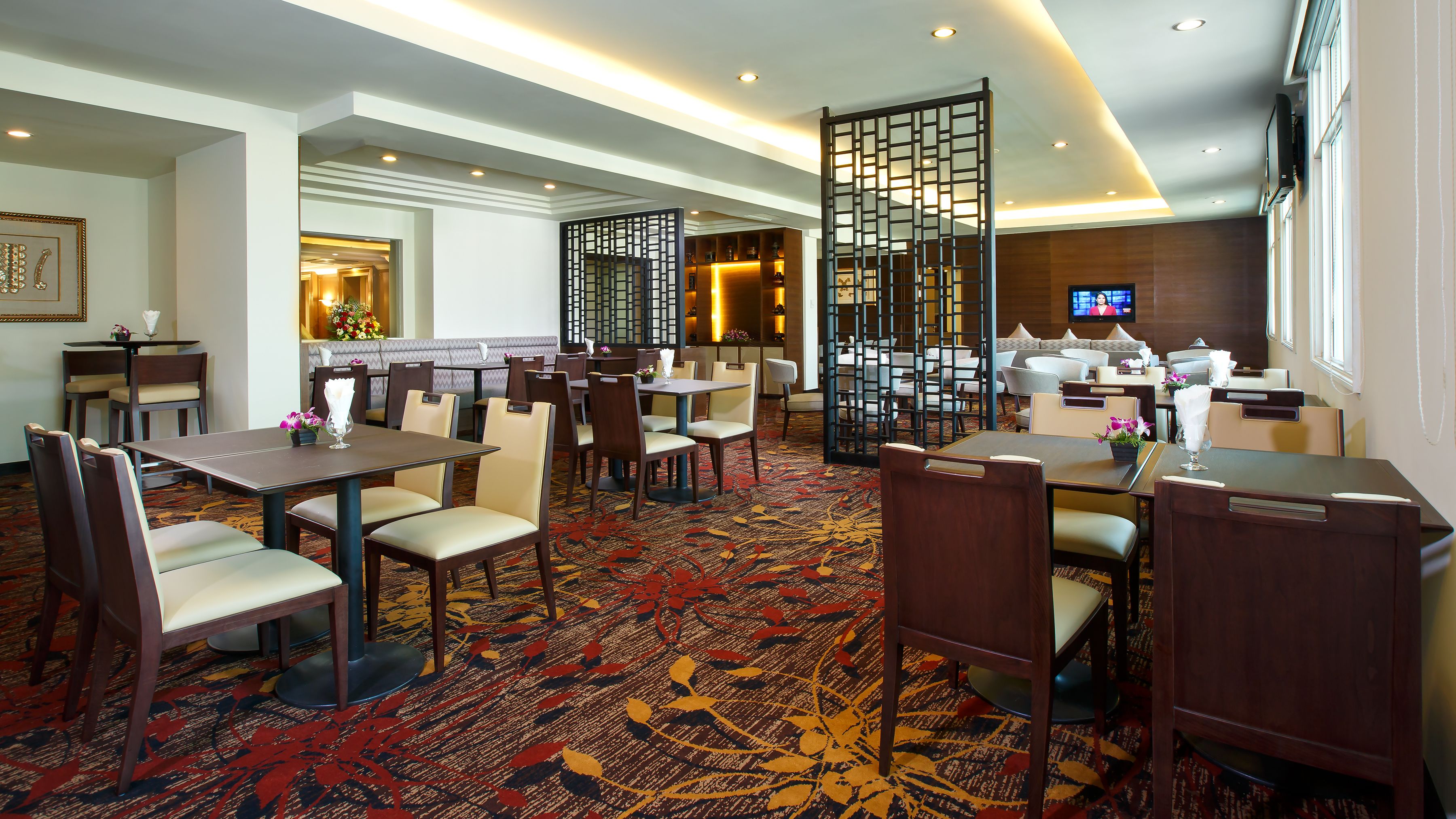 A hotel dining area with multiple tables and chairs, a patterned carpet, and a television screen.