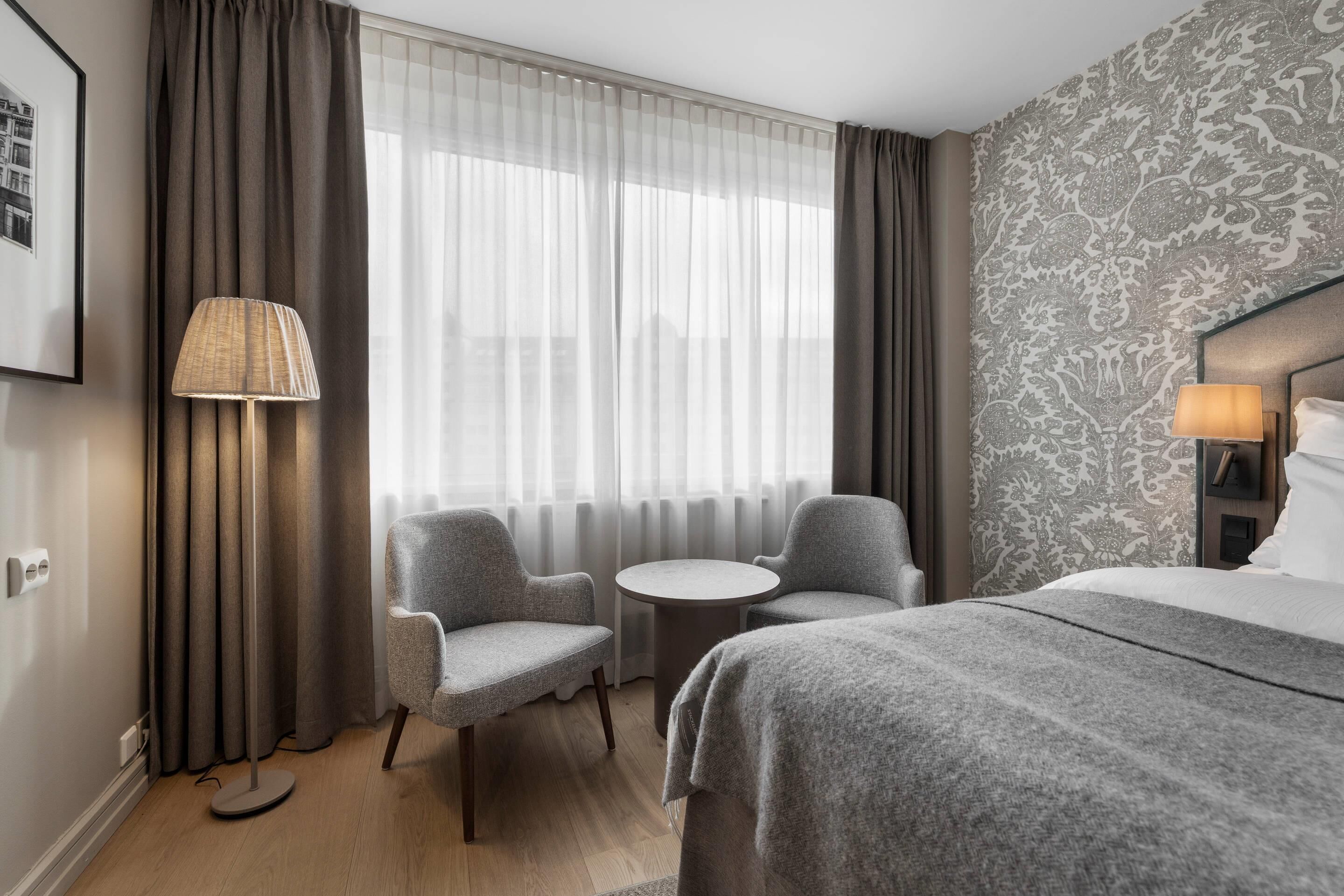 A hotel room with a bed, two armchairs, a small table, a lamp, and a view of the city through sheer curtains.