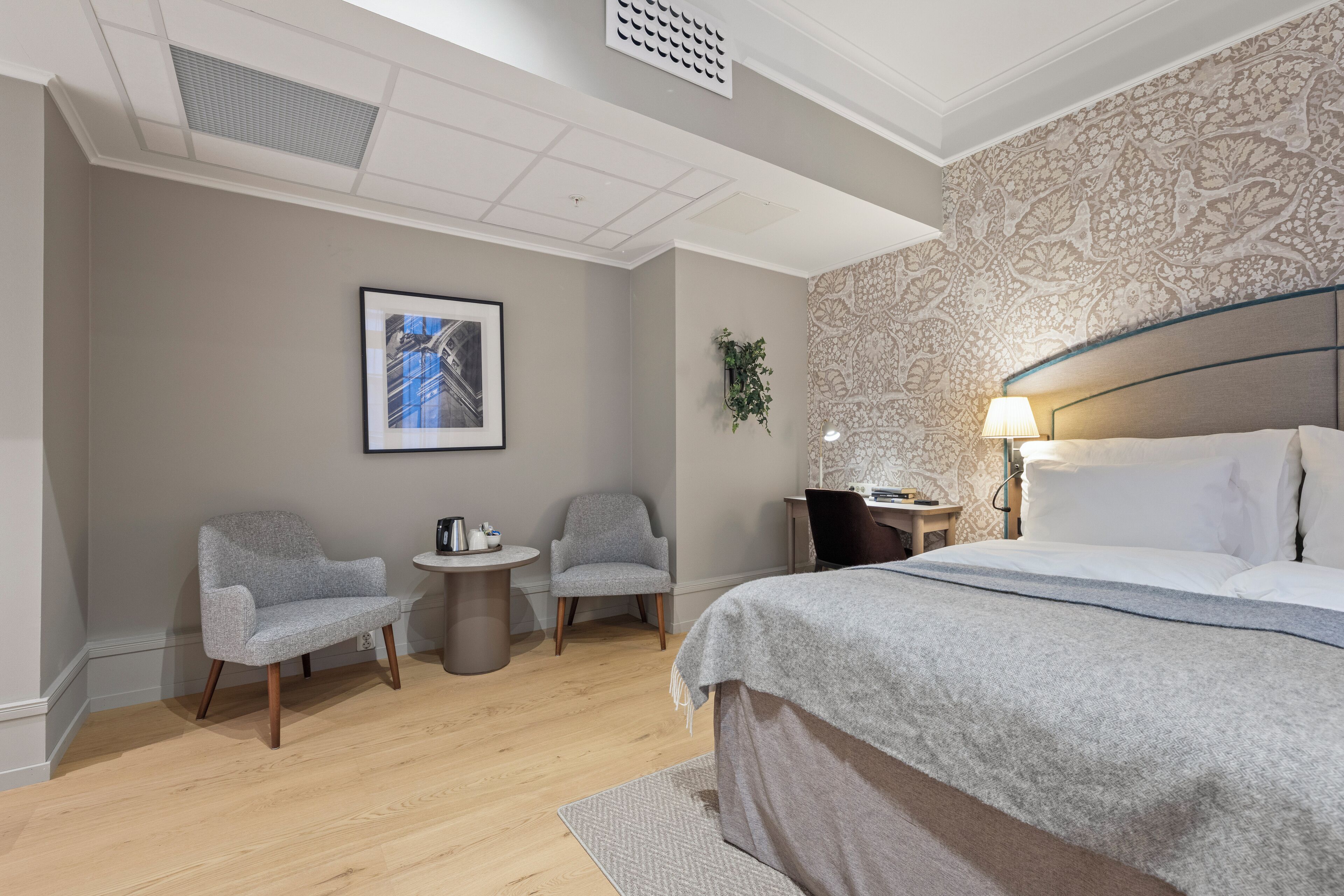 A modern hotel room with a large bed, two armchairs, a small round table, and a wall-mounted plant.