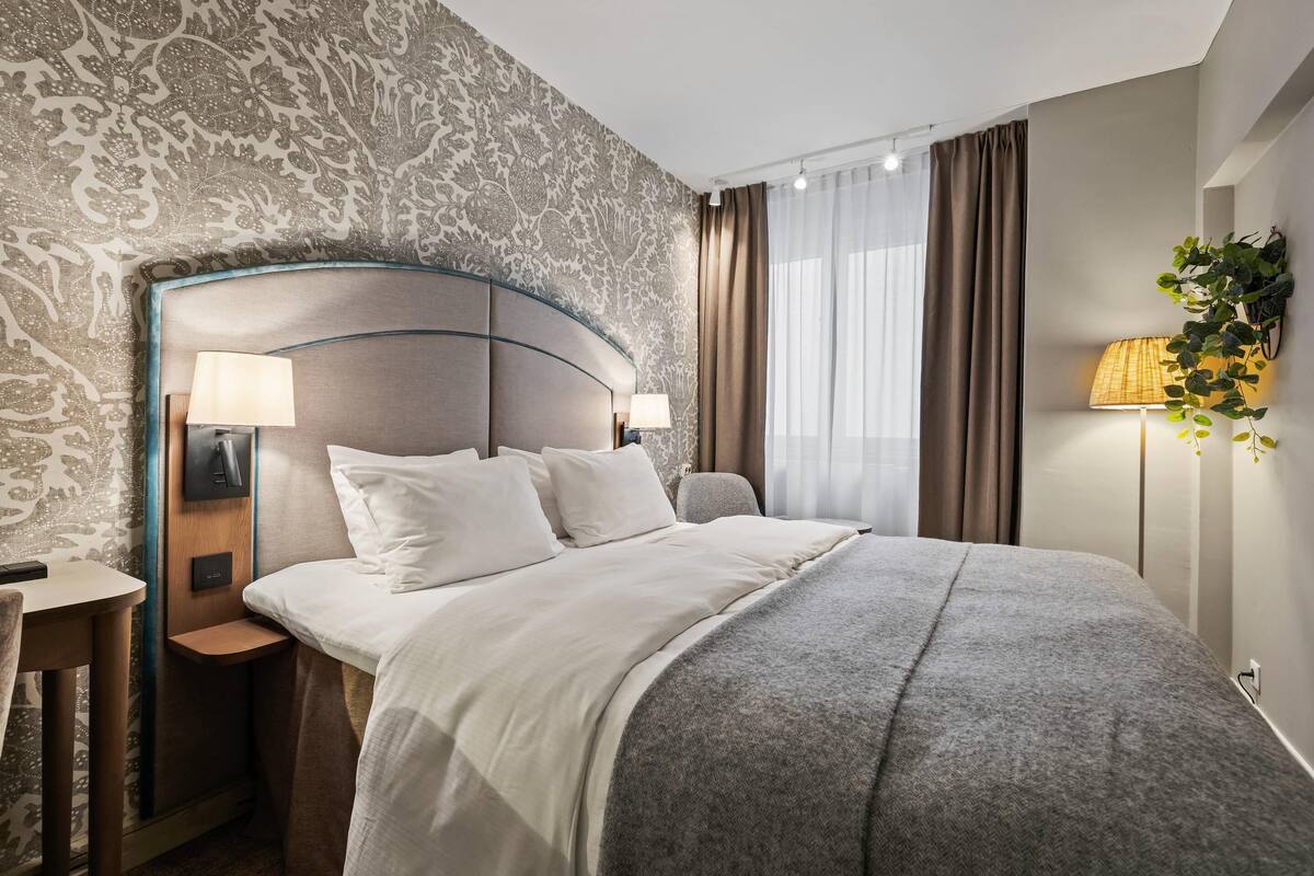 Habitación de hotel ordenada con una cama grande, dos mesitas de noche con lámparas, una pequeña mesa y una planta en la pared.