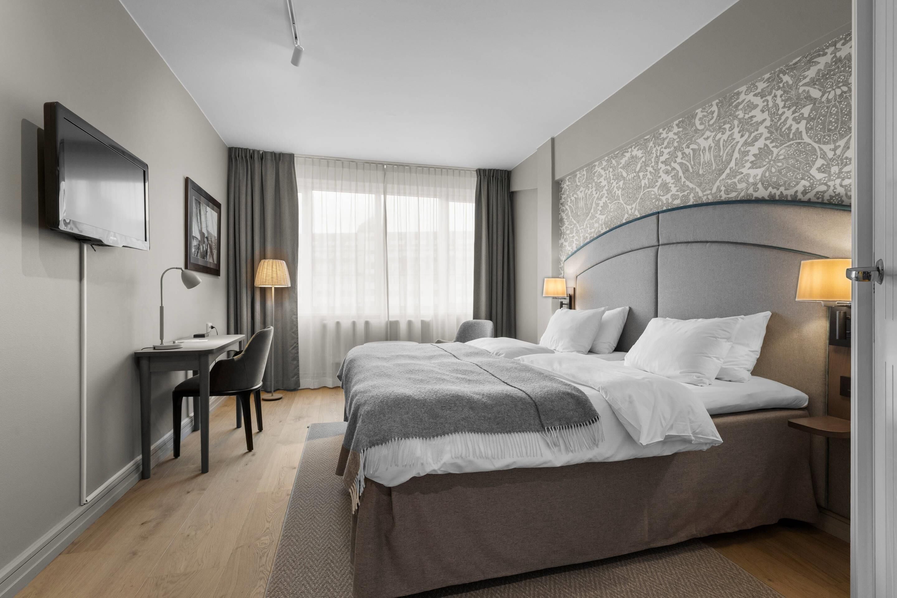 Una habitació d'hotel moderna amb un llit gran, dues taules de nit amb làmpades, un escriptori amb una cadira i una televisió penjada a la paret.