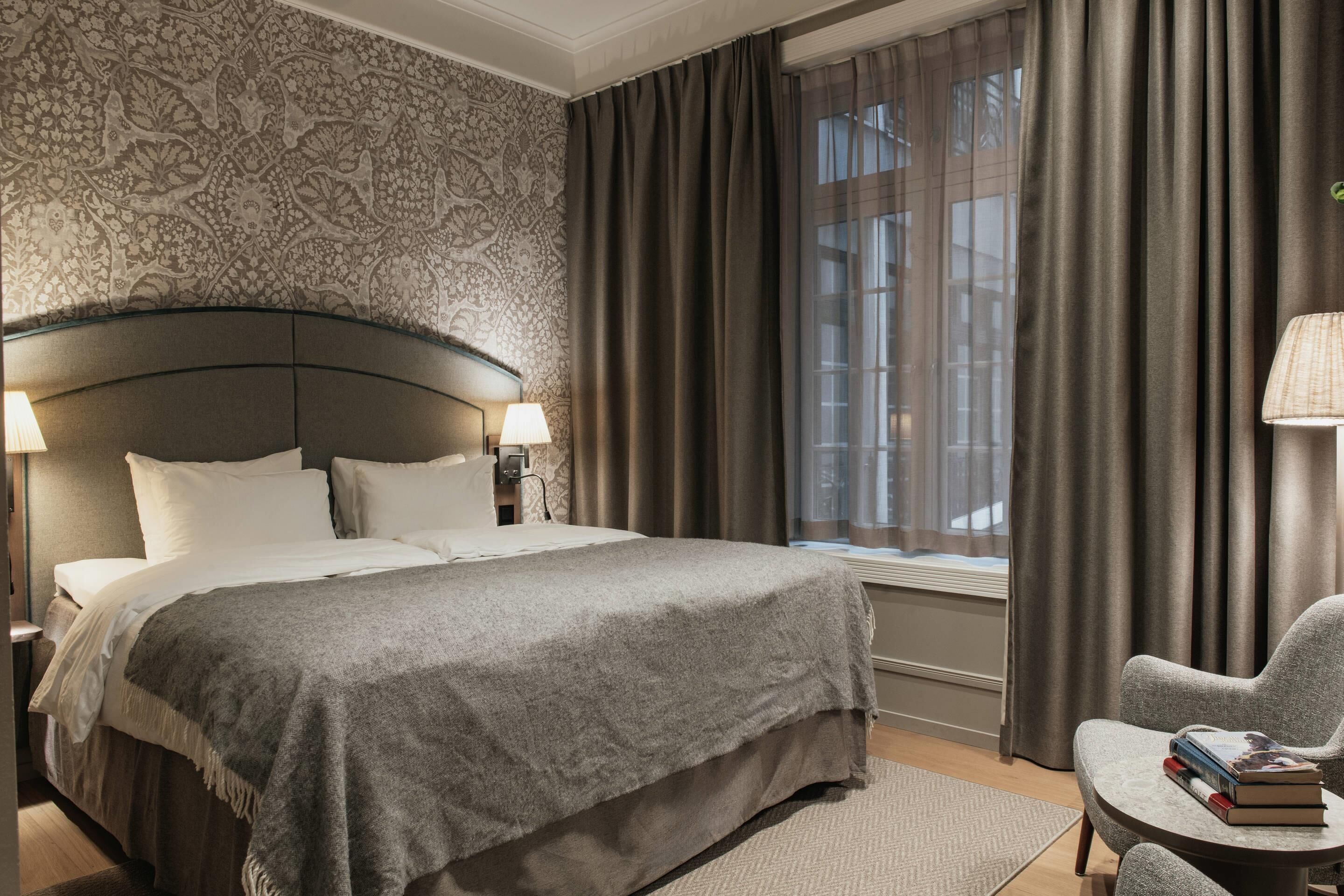 A hotel room with a large bed, a chair, a small table, and a window with curtains.