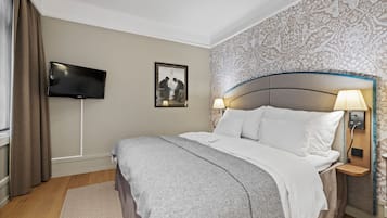 A neatly arranged hotel room with a large bed, a flat-screen TV mounted on the wall, a bedside table with a lamp, and a patterned wallpaper.