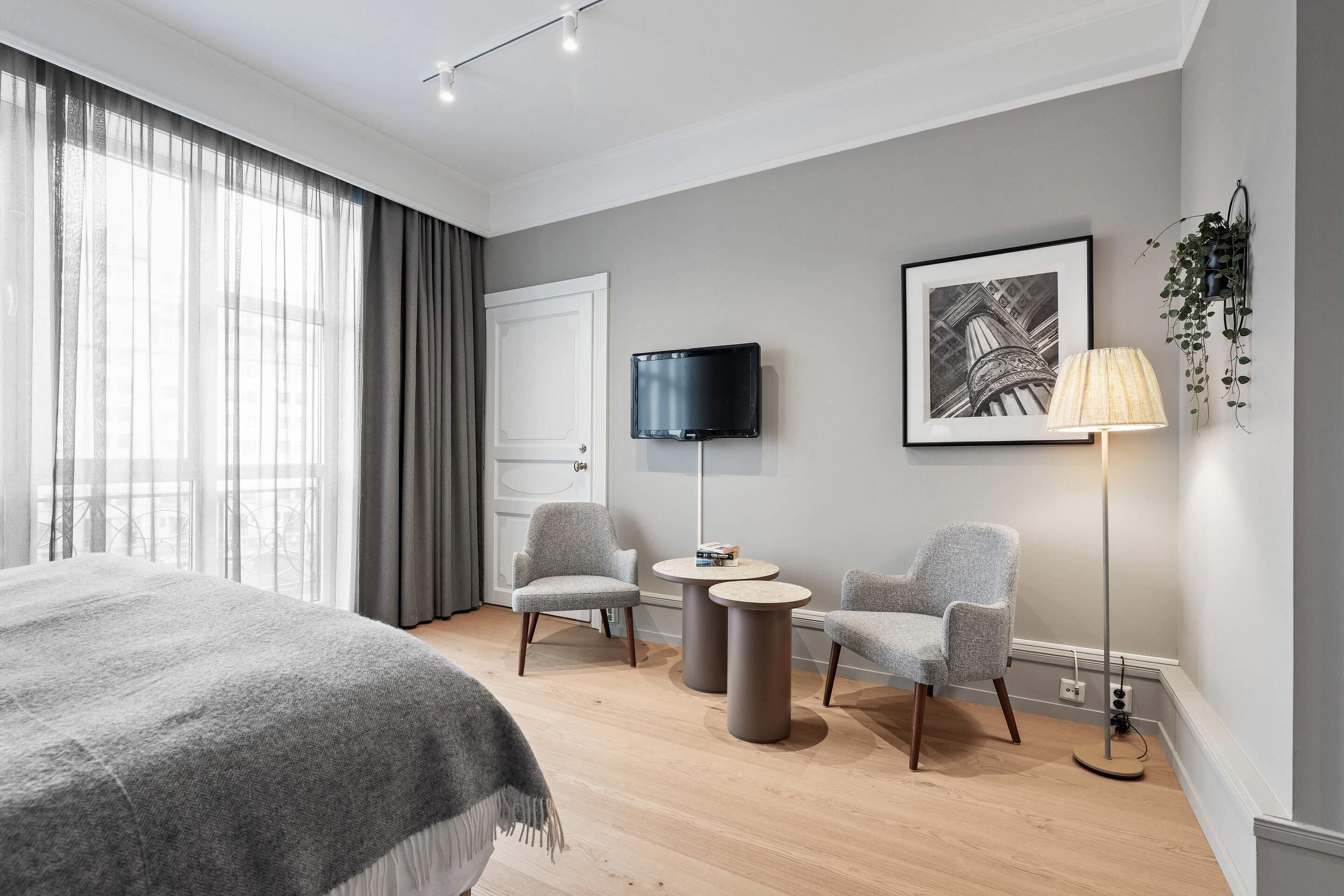 A modern hotel room with a large bed, two armchairs, a small table, a TV, and a framed picture on the wall.