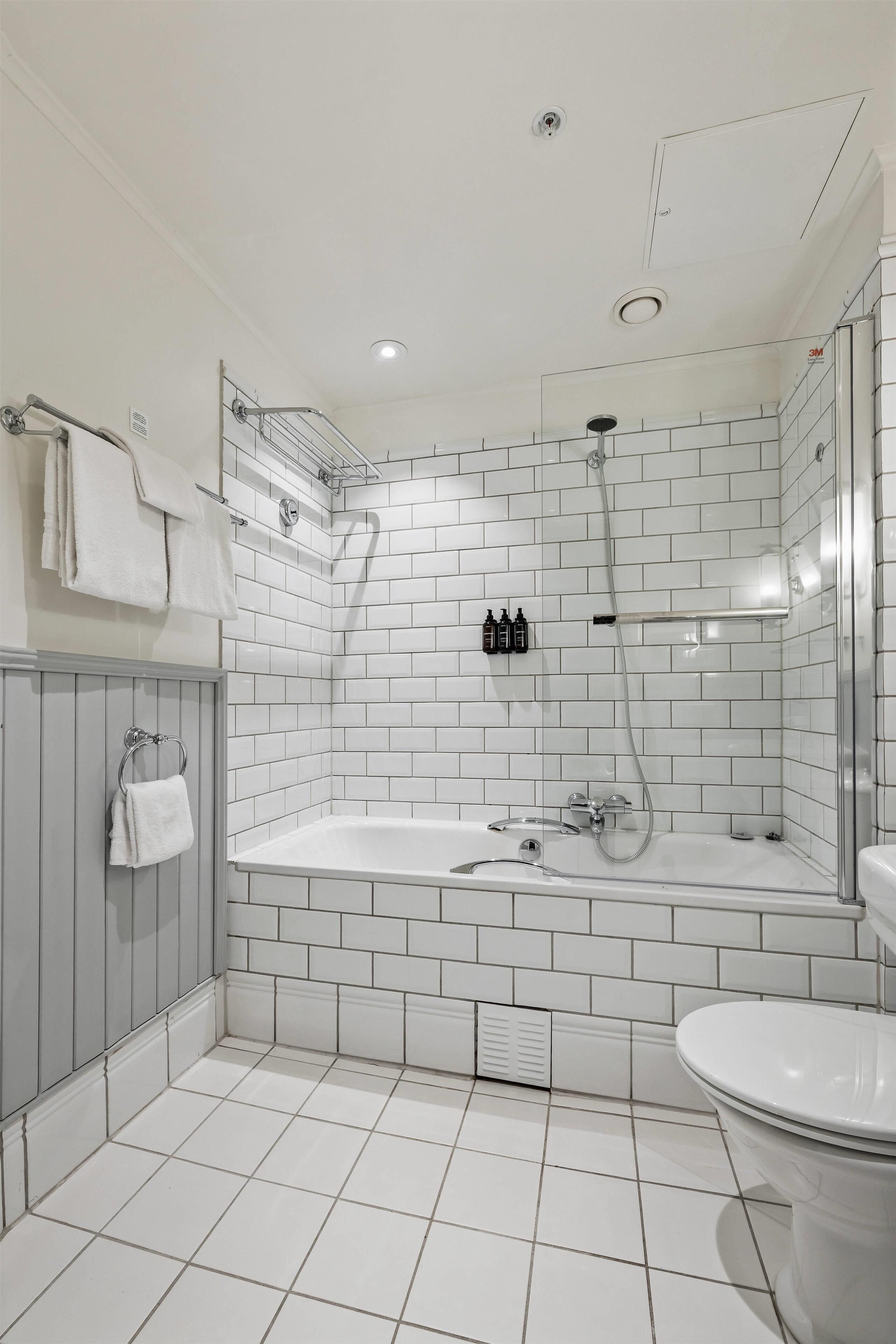 A modern bathroom with white tiled walls and floor, a glass-enclosed shower, and a white bathtub.