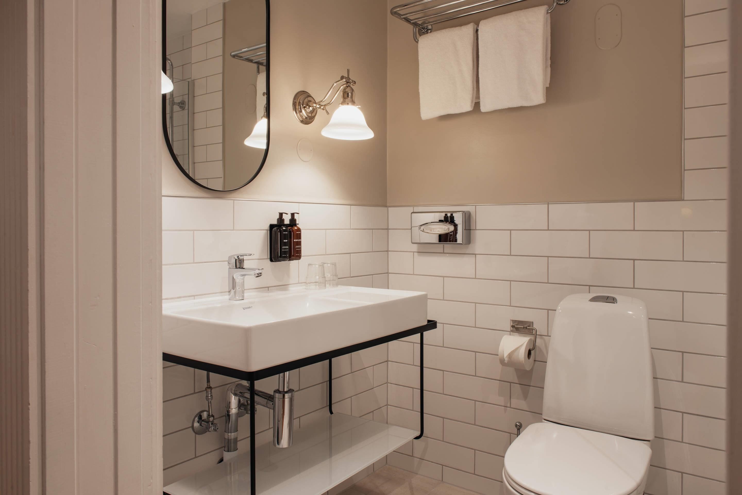 A modern bathroom with a white sink, mirror, and toilet. There are two wall-mounted lights, a towel rack, and a small shelf with toiletries.