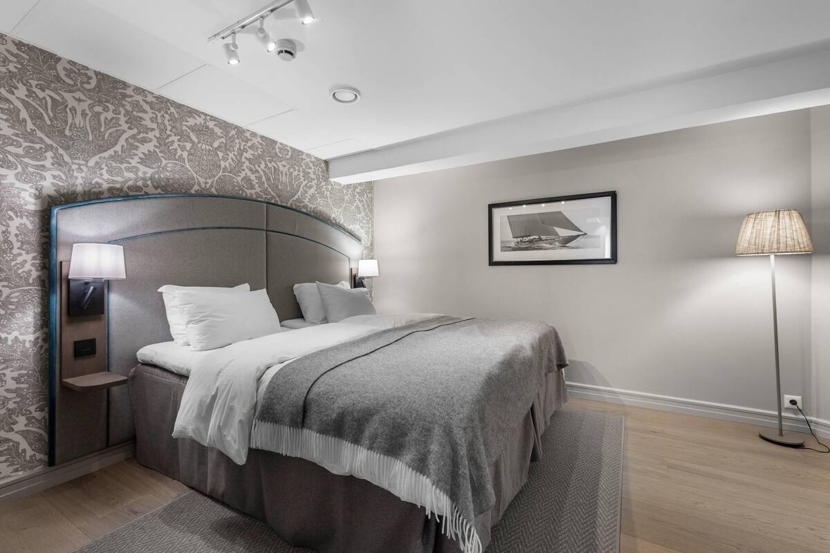 Un dormitorio moderno con cabecera curvada, una mesita de noche con lámpara, cama con manta gris, lámpara de pie y un cuadro enmarcado en la pared.