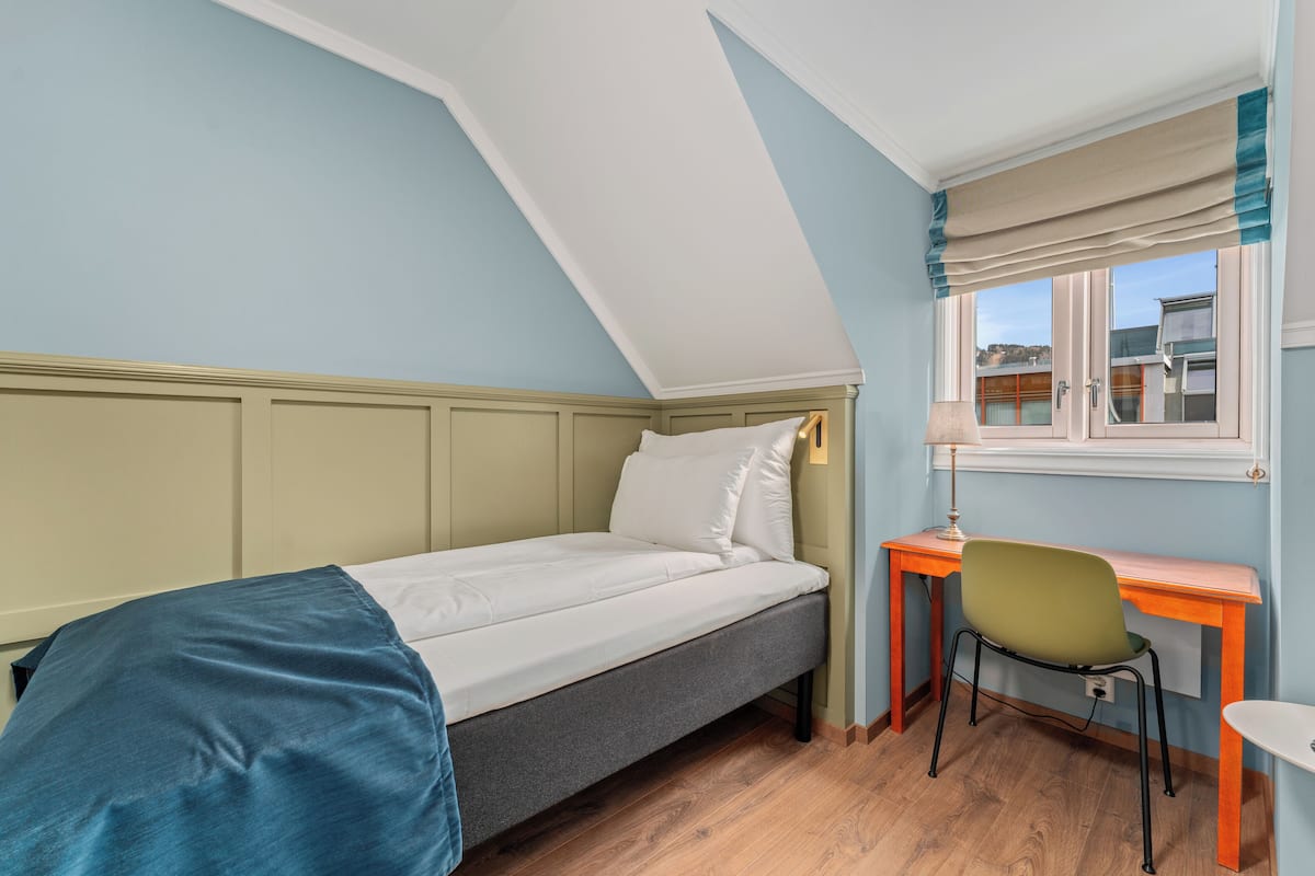 A single bed with a blue blanket, a desk with a chair, and a window with curtains.