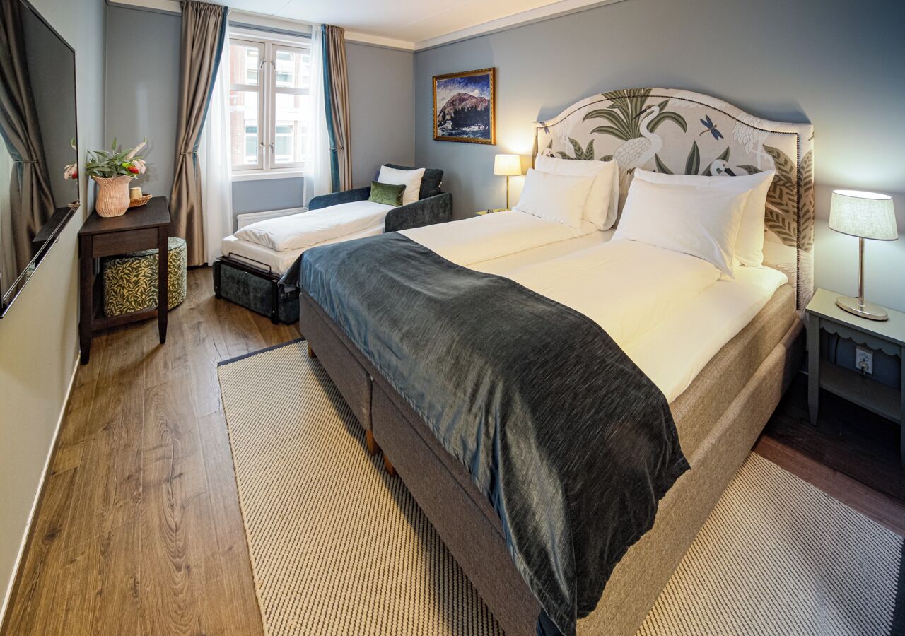 A hotel room with a large bed, a smaller bed, a nightstand, a lamp, a painting, and a window with curtains.