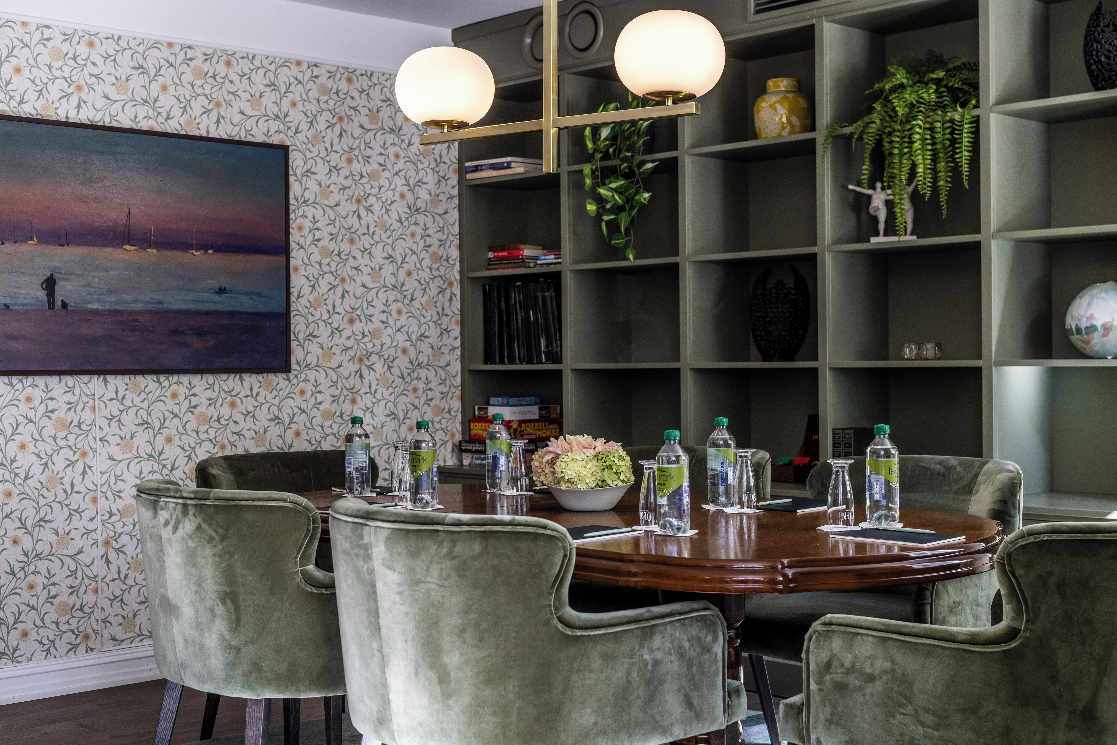 Un comedor con una mesa redonda, sillas tapizadas en verde y una estantería con diversos objetos.