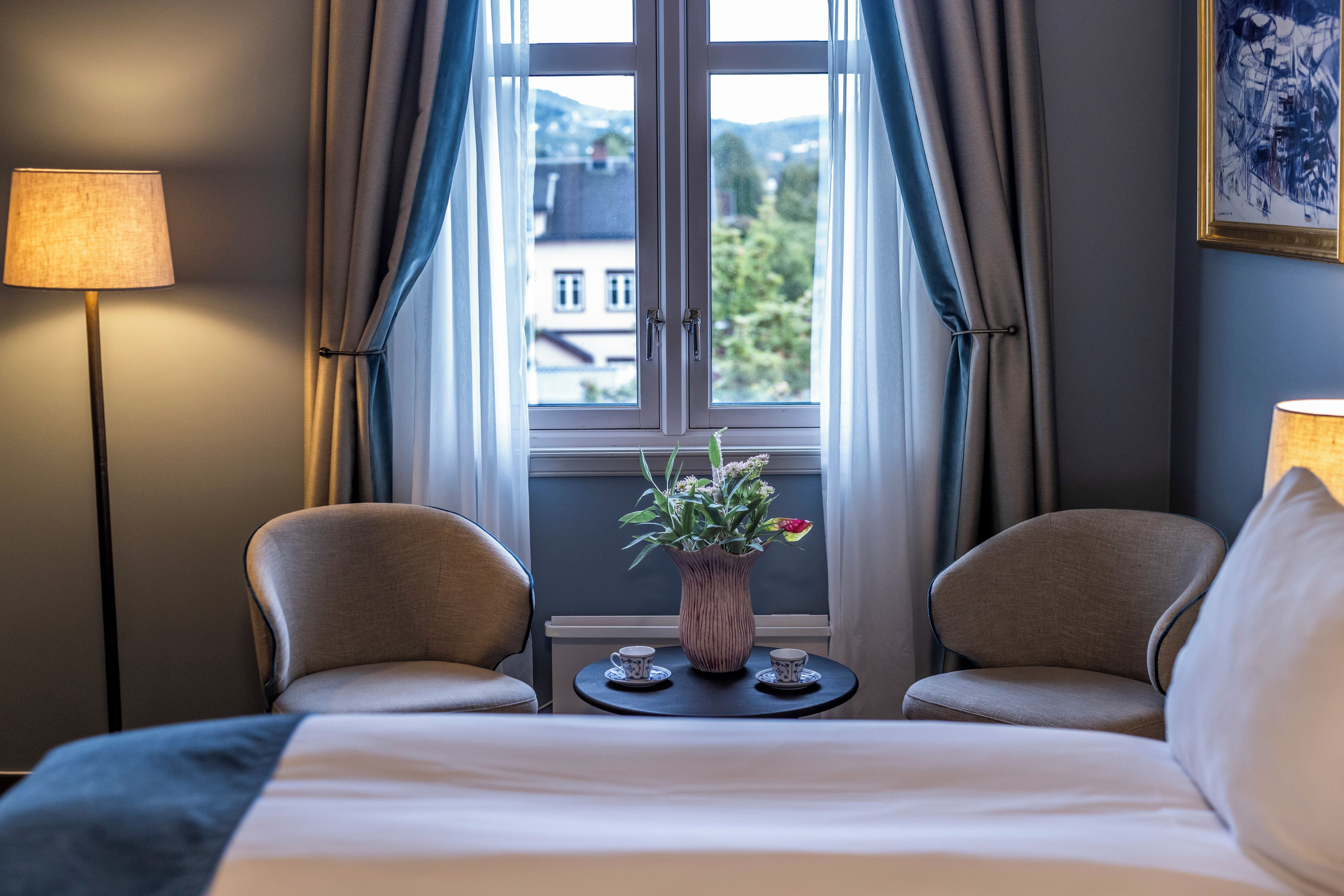 A hotel room with a bed, two chairs, a small table with a vase of flowers, and a window with curtains.